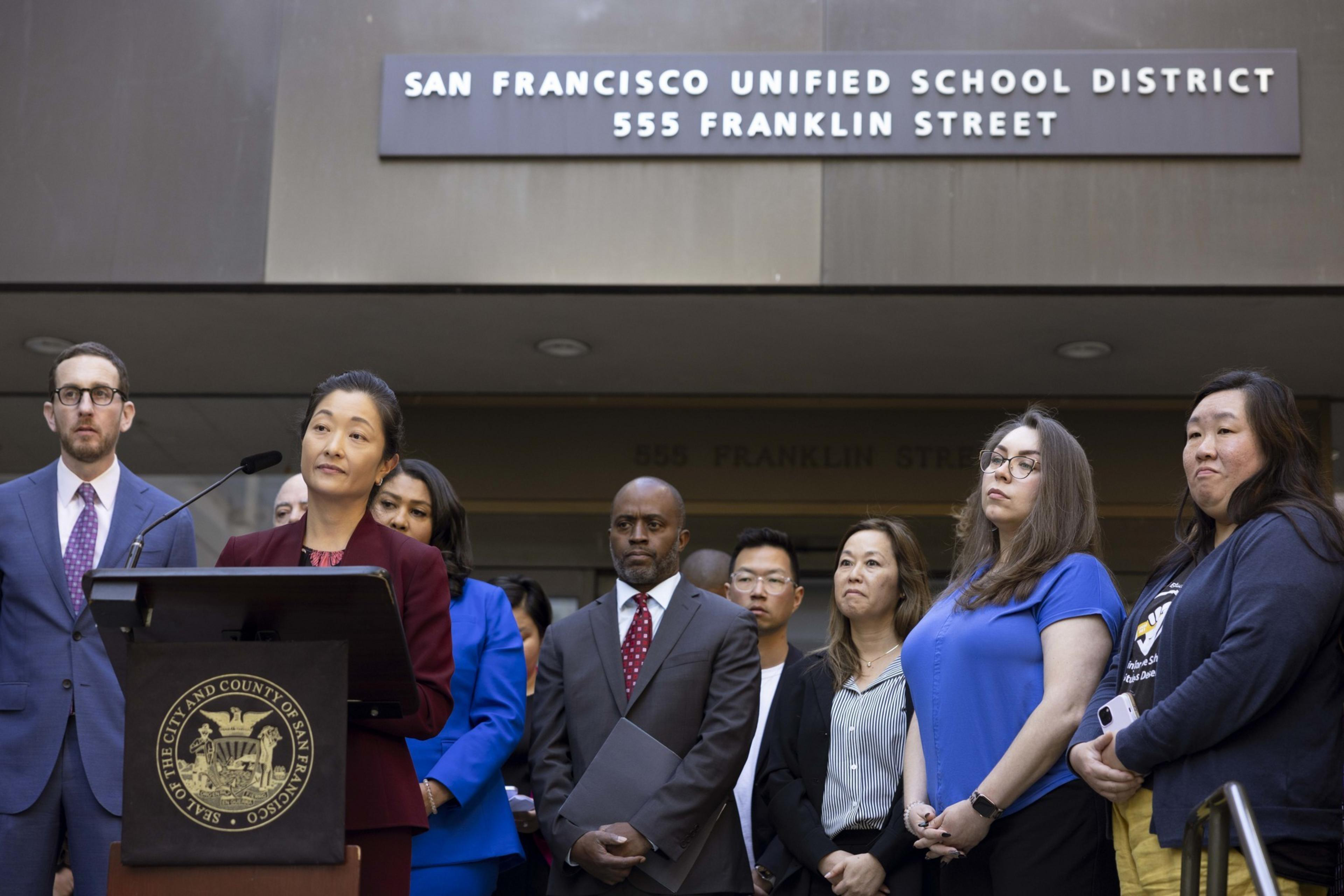 A group of individuals stands outside the San Francisco Unified School District building. A woman at a podium is speaking, surrounded by attentive people.
