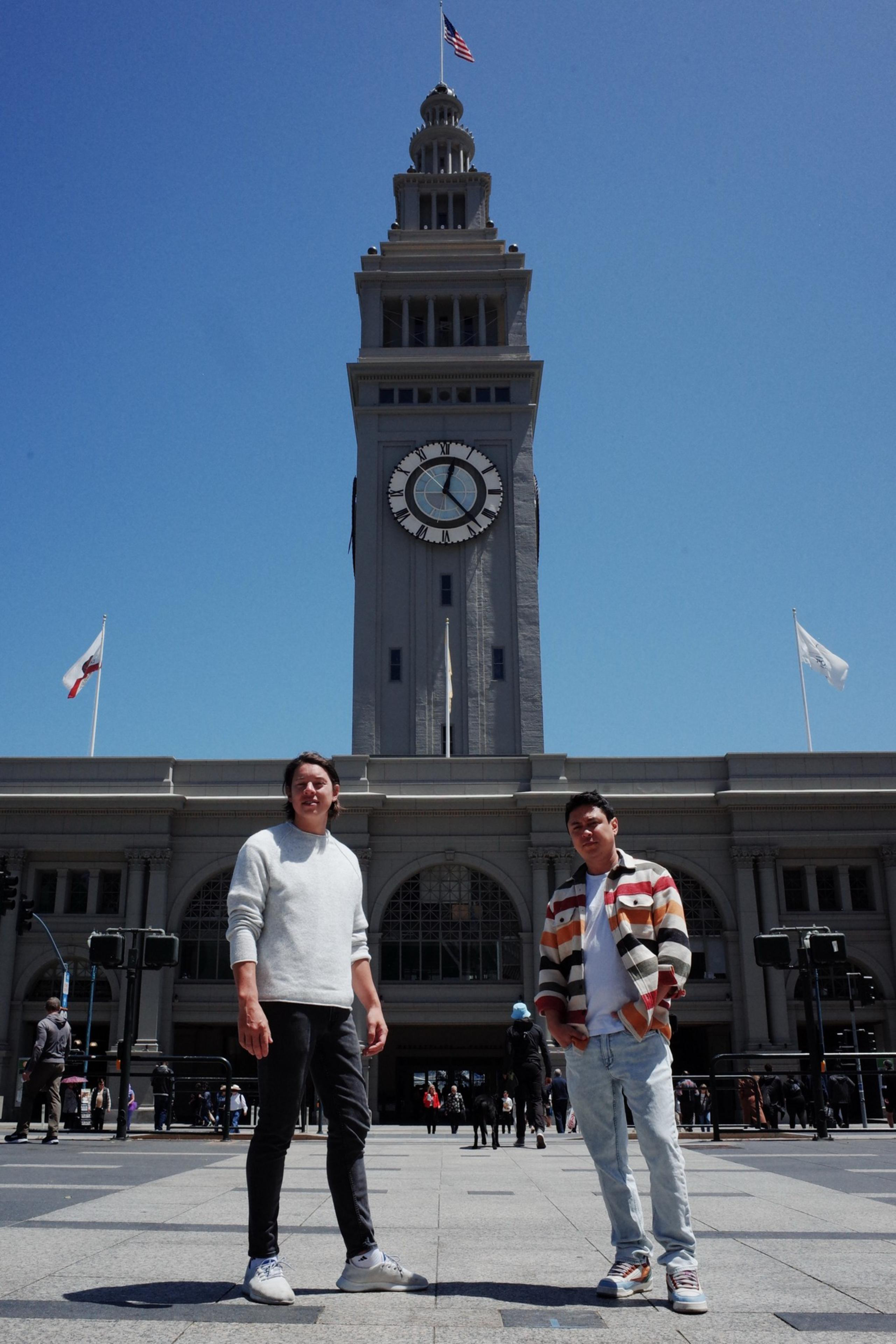 Two people stand in front of a large clock tower with arched windows. The sky is clear and blue, and flags are flying atop the building.