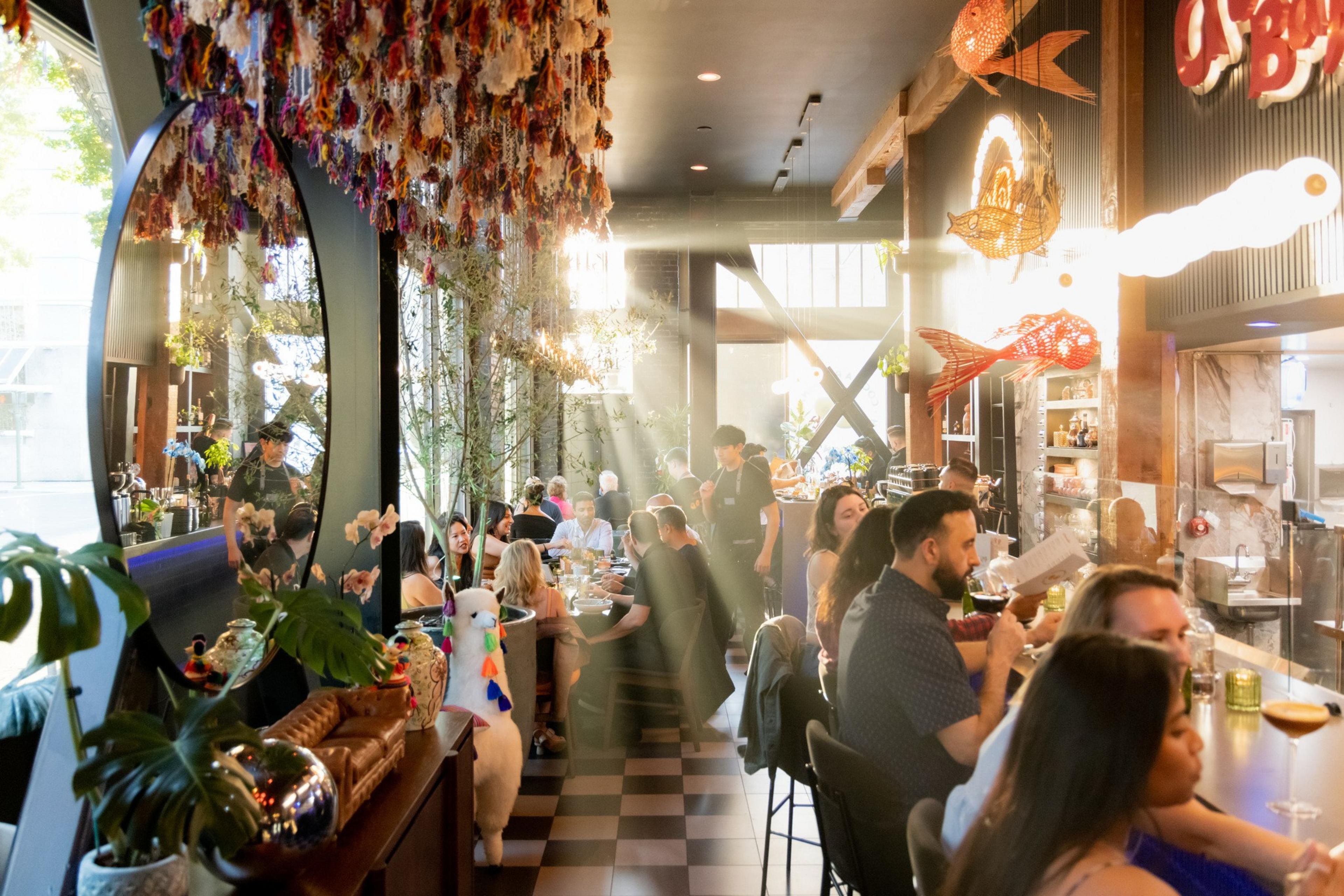 The image shows a bustling restaurant with people dining. Sunlight streams through large windows, illuminating hanging decorations and a lively atmosphere.