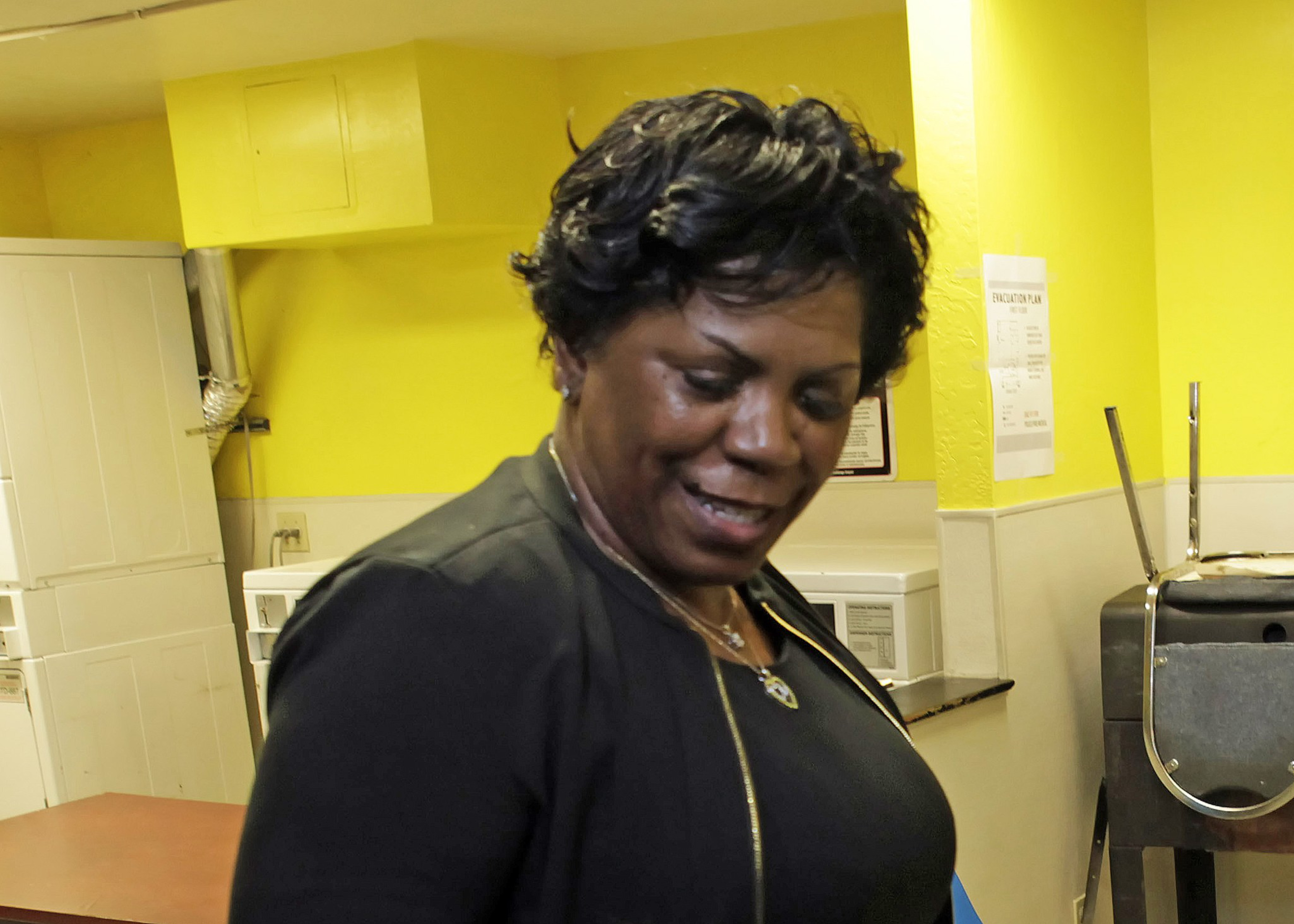 A woman with short, curly black hair smiles while looking down, wearing a black jacket and necklace; the background is a yellow-painted room.