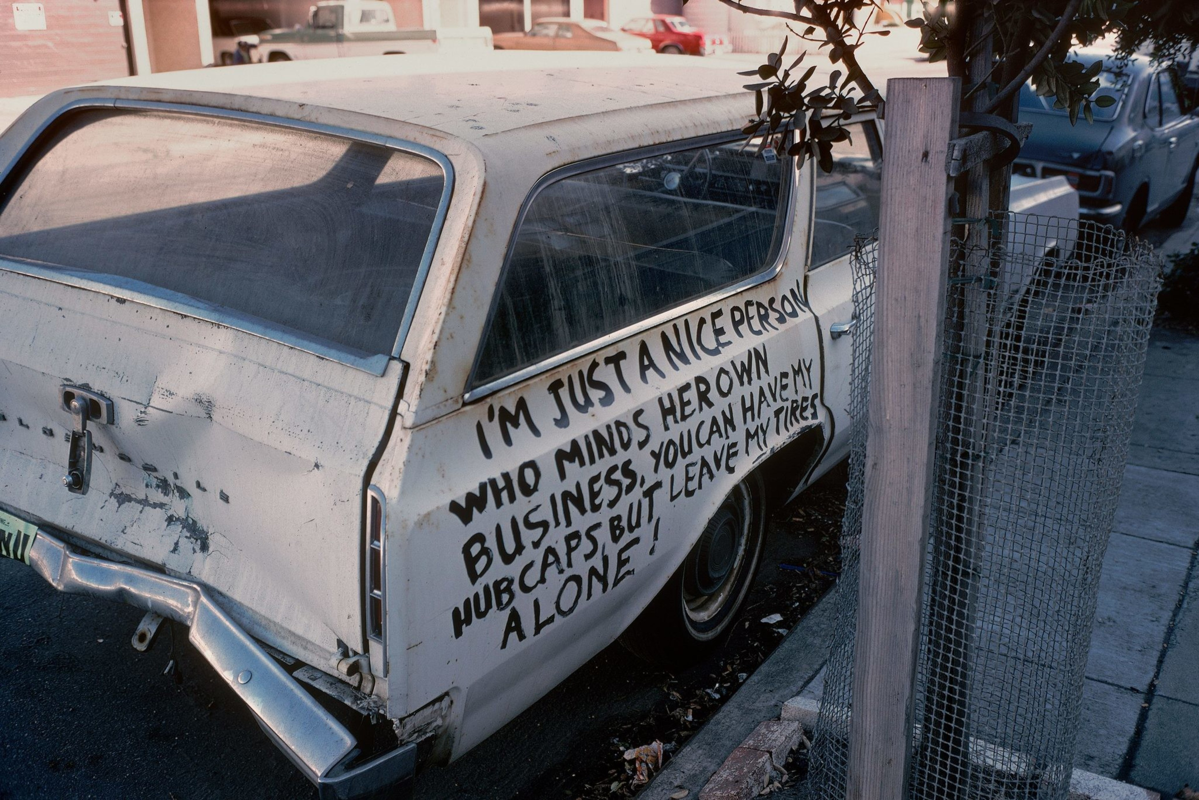 A white car is parked with a handwritten message on its side saying, &quot;I'm just a nice person who minds her own business. You can have my hubcaps, but leave my tires alone!&quot;