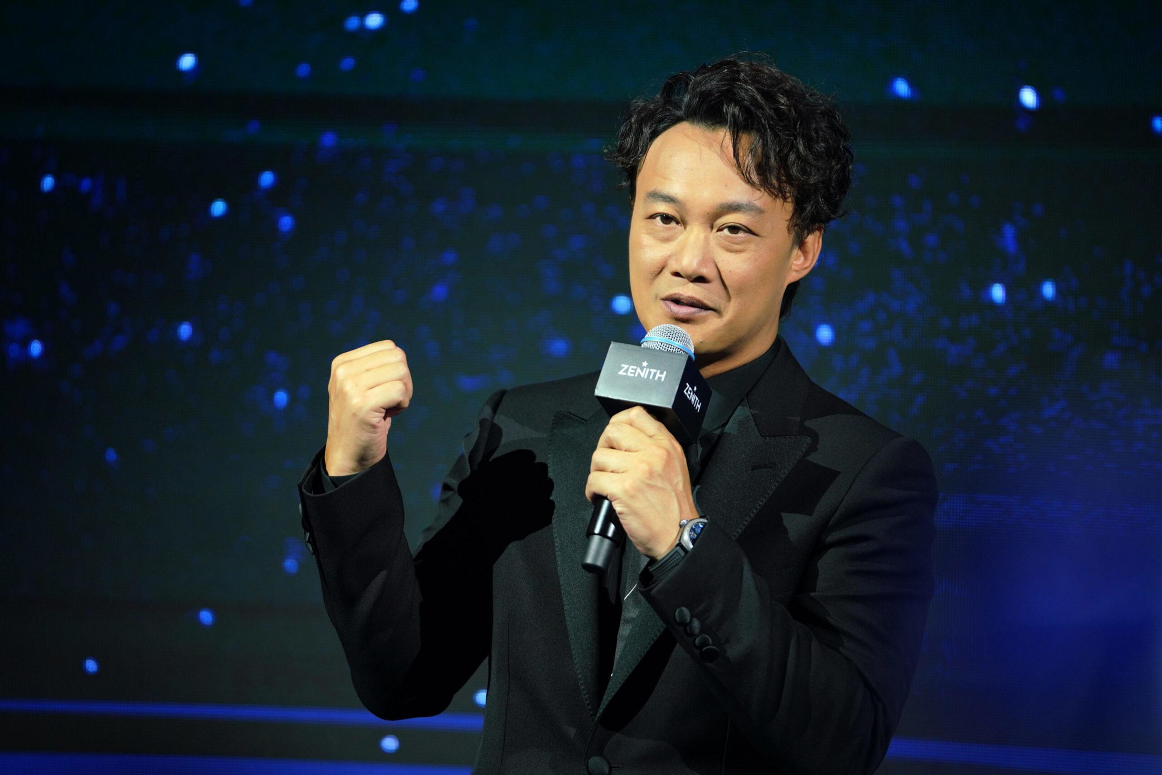 A man with curly hair wearing a black suit holds a microphone in one hand and raises his other fist, standing against a dark blue starry background.