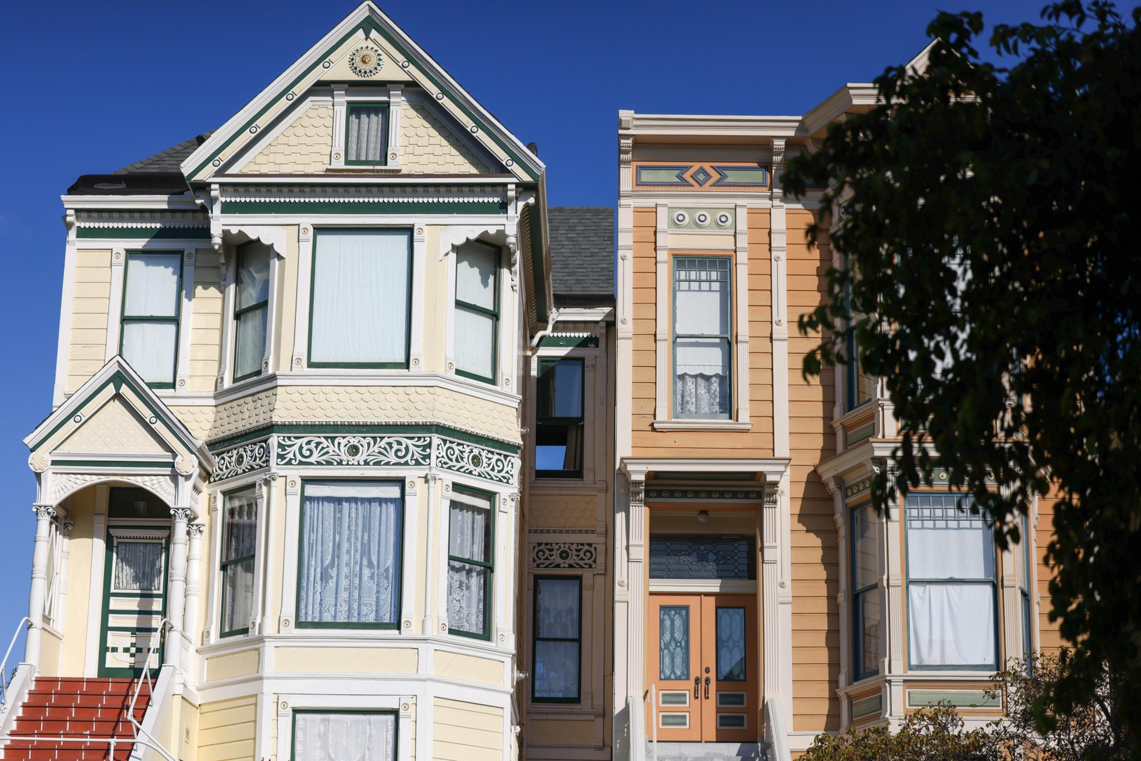 Two adjacent Victorian-style houses with ornate trim, large windows with lace curtains, and warm pastel colors under a clear blue sky.
