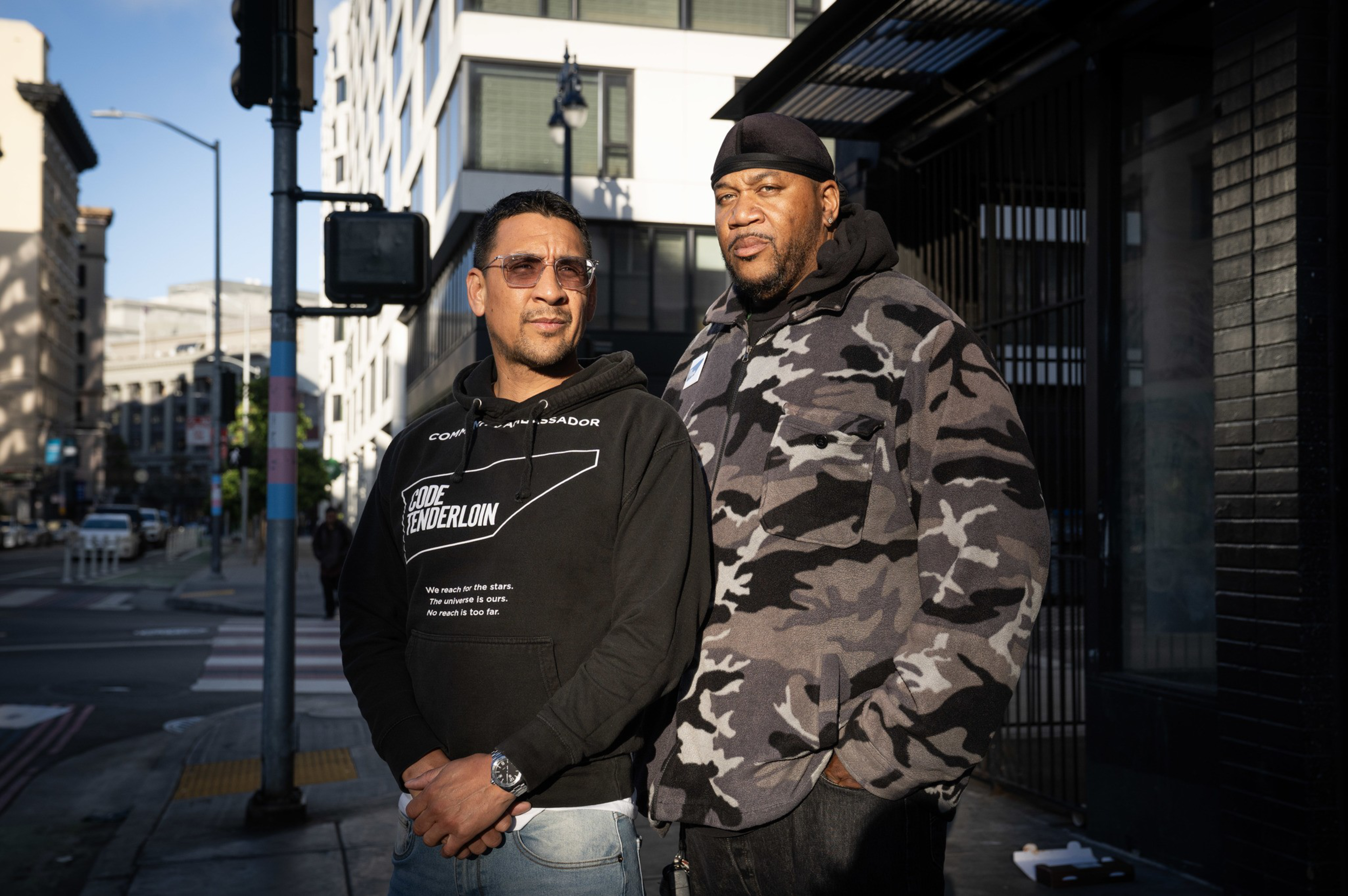 Two men stand side by side on a city sidewalk, one wearing sunglasses and a black hoodie, the other in a gray camouflage jacket and black head covering.