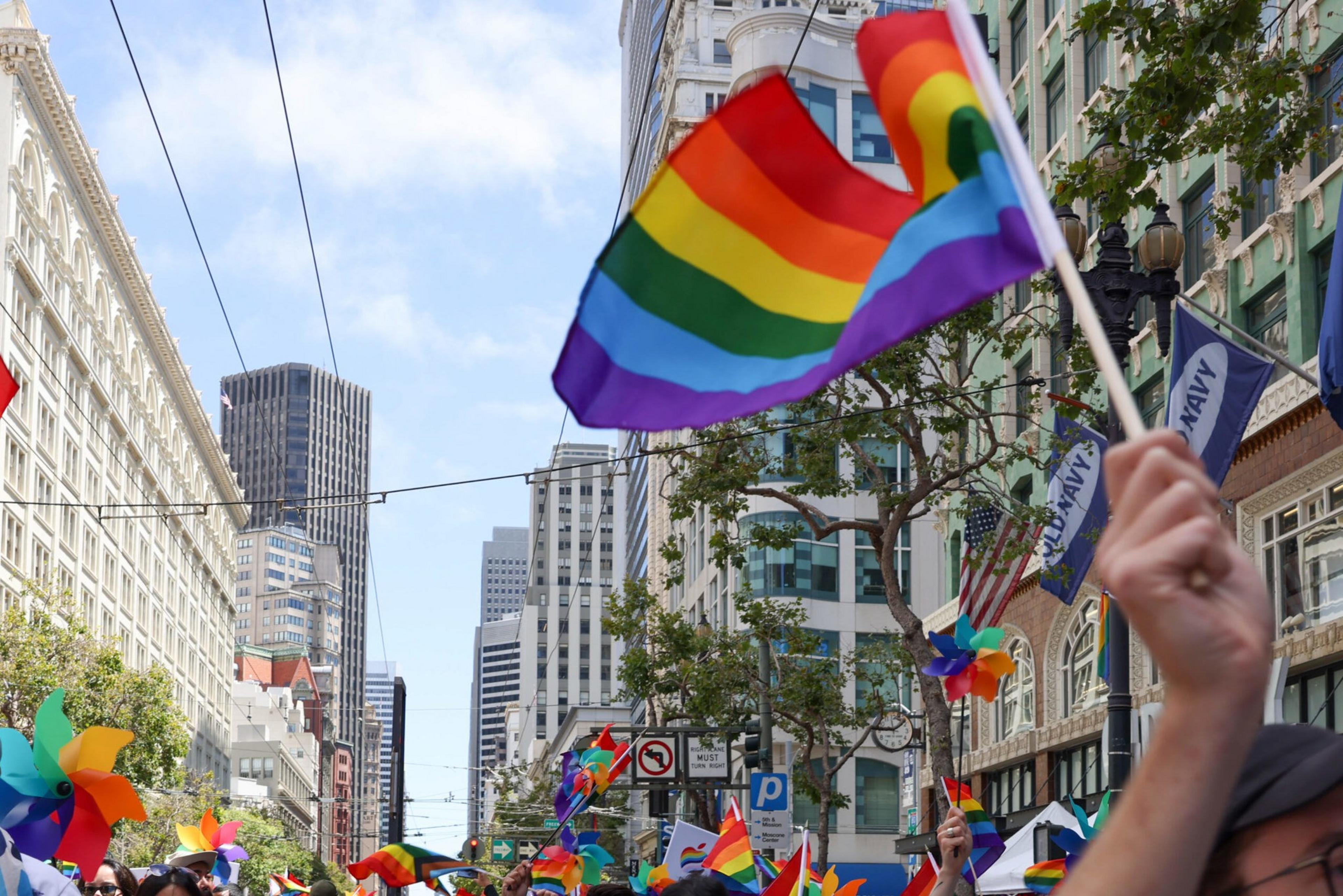 A rainbow pride flag waves above a crowd on a city street lined with tall buildings and colorful pinwheels, under a partly cloudy blue sky.