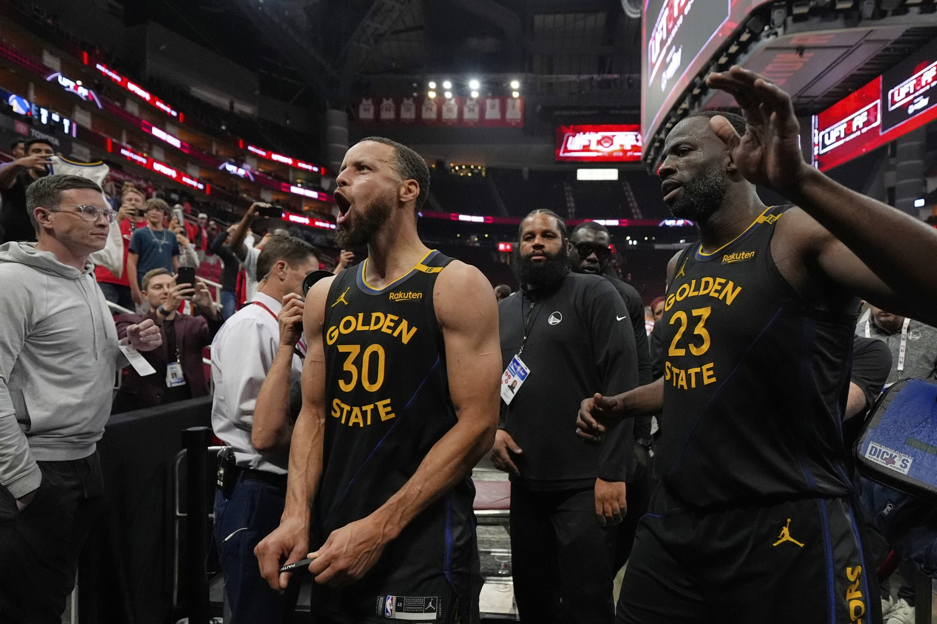 Two basketball players in Golden State jerseys, numbered 30 and 23, walk off the court, with one looking energized. Fans and staff surround them, taking photos.