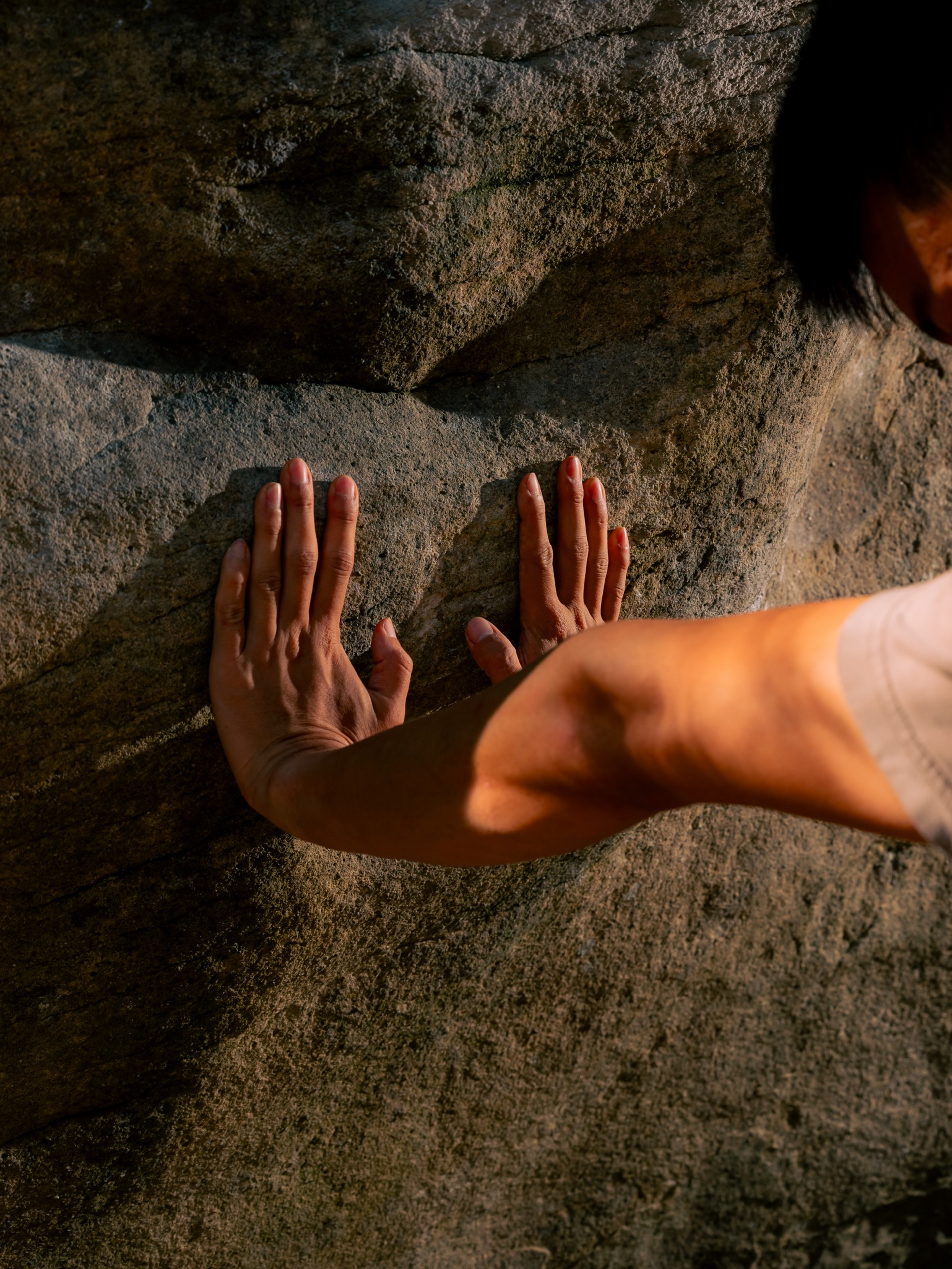 Two hands pressing flat against a textured rock surface, with part of a forearm and shoulder visible, illuminated by warm light.