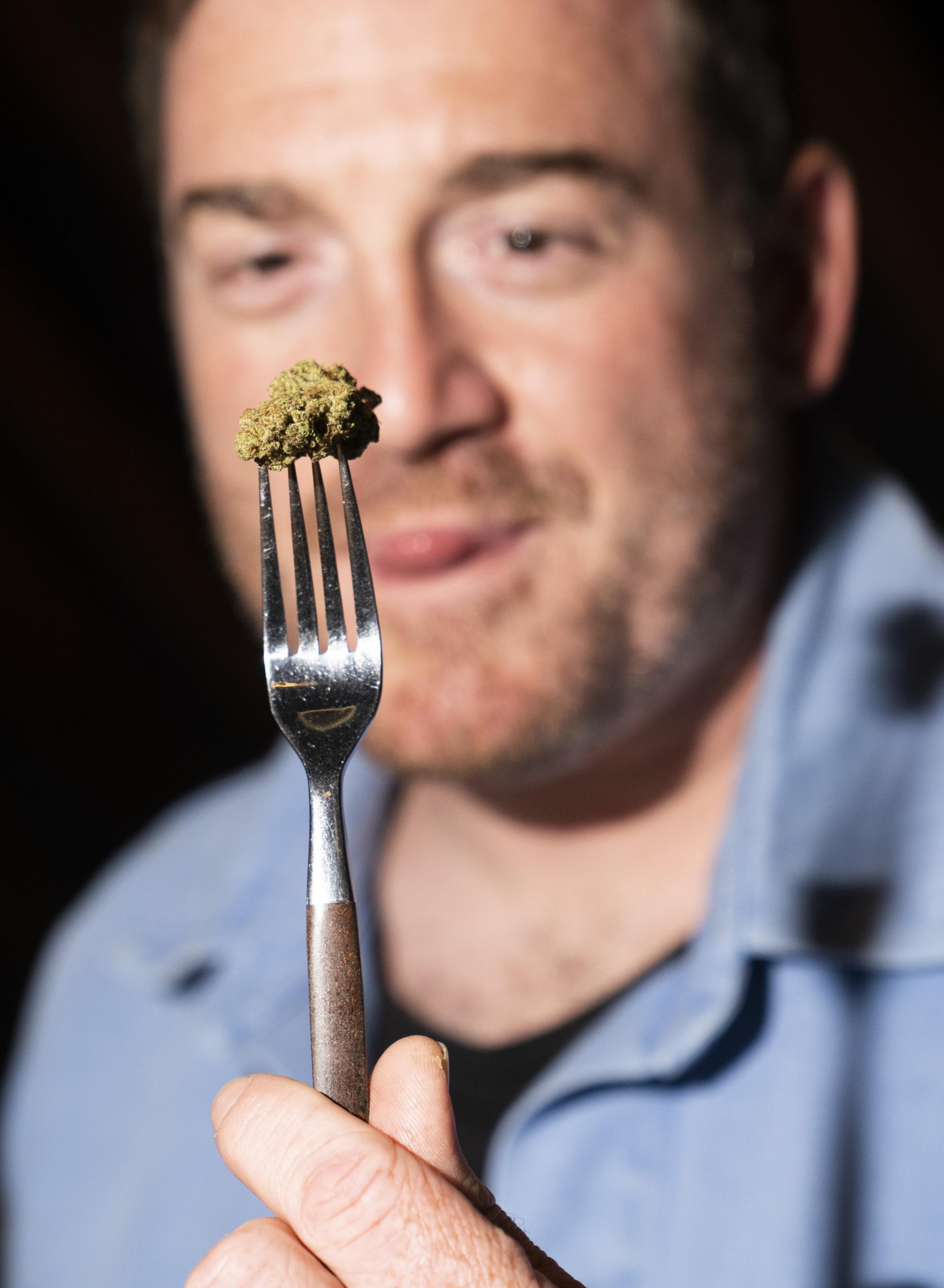 A man holding a fork with a small, greenish-brown piece of food or substance on it, focusing on the fork while smiling slightly in a blurred background.