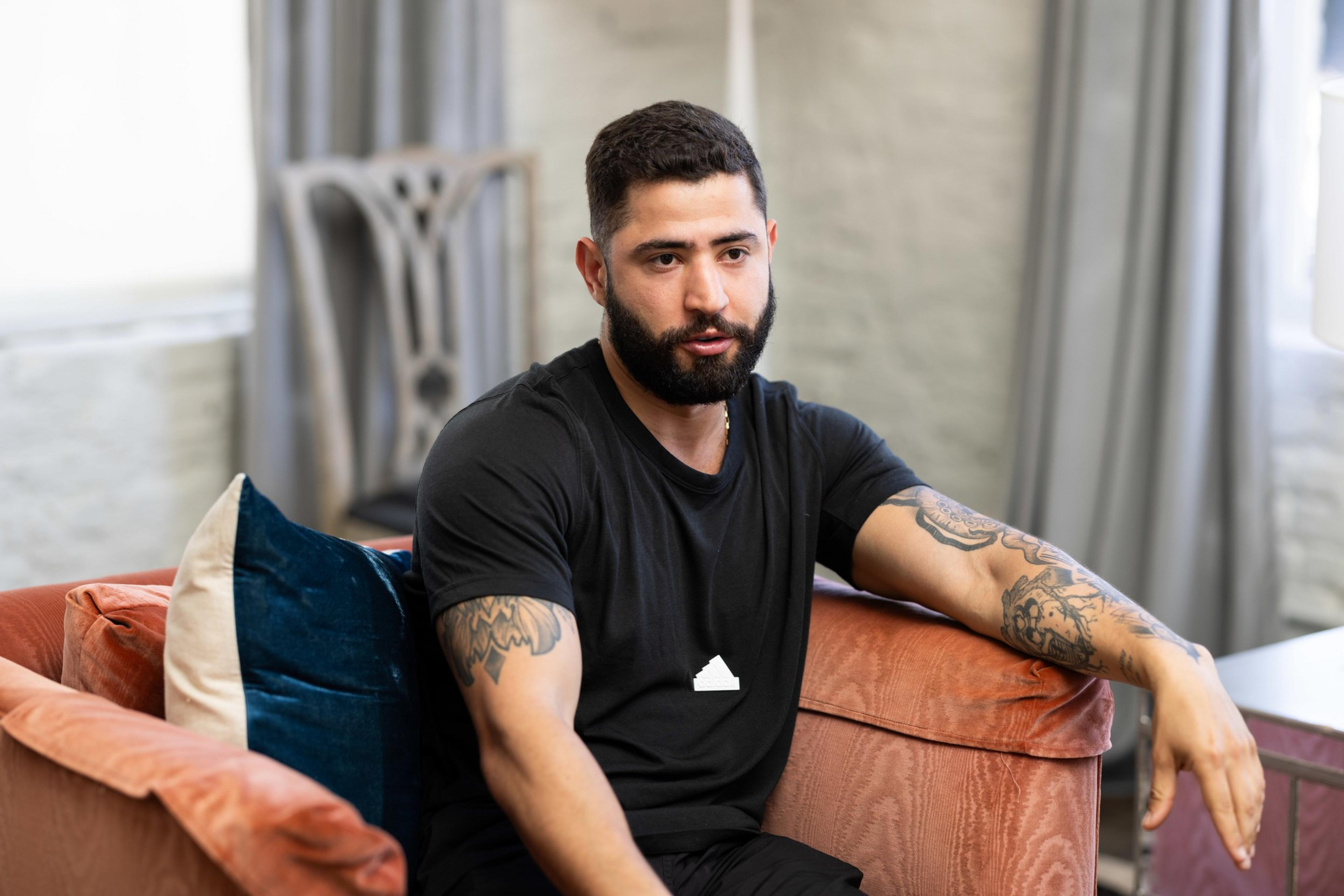 A bearded man with tattoos on both arms is sitting on an orange couch, dressed in a black shirt. He appears to be in a casual setting with a chair and curtains in the background.