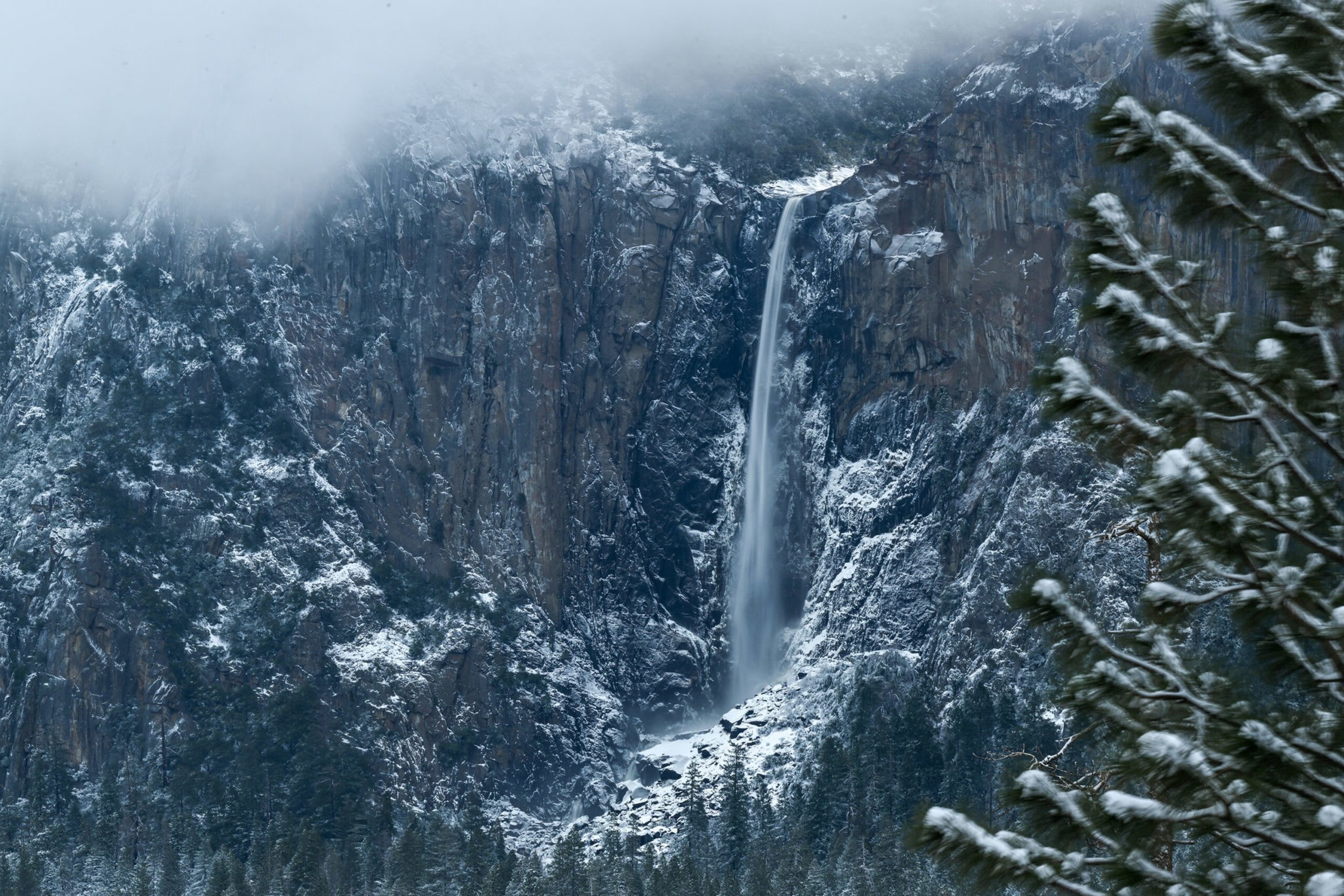 A tall waterfall cascades down a snow-dusted rocky cliff surrounded by evergreen trees under a cloudy sky.