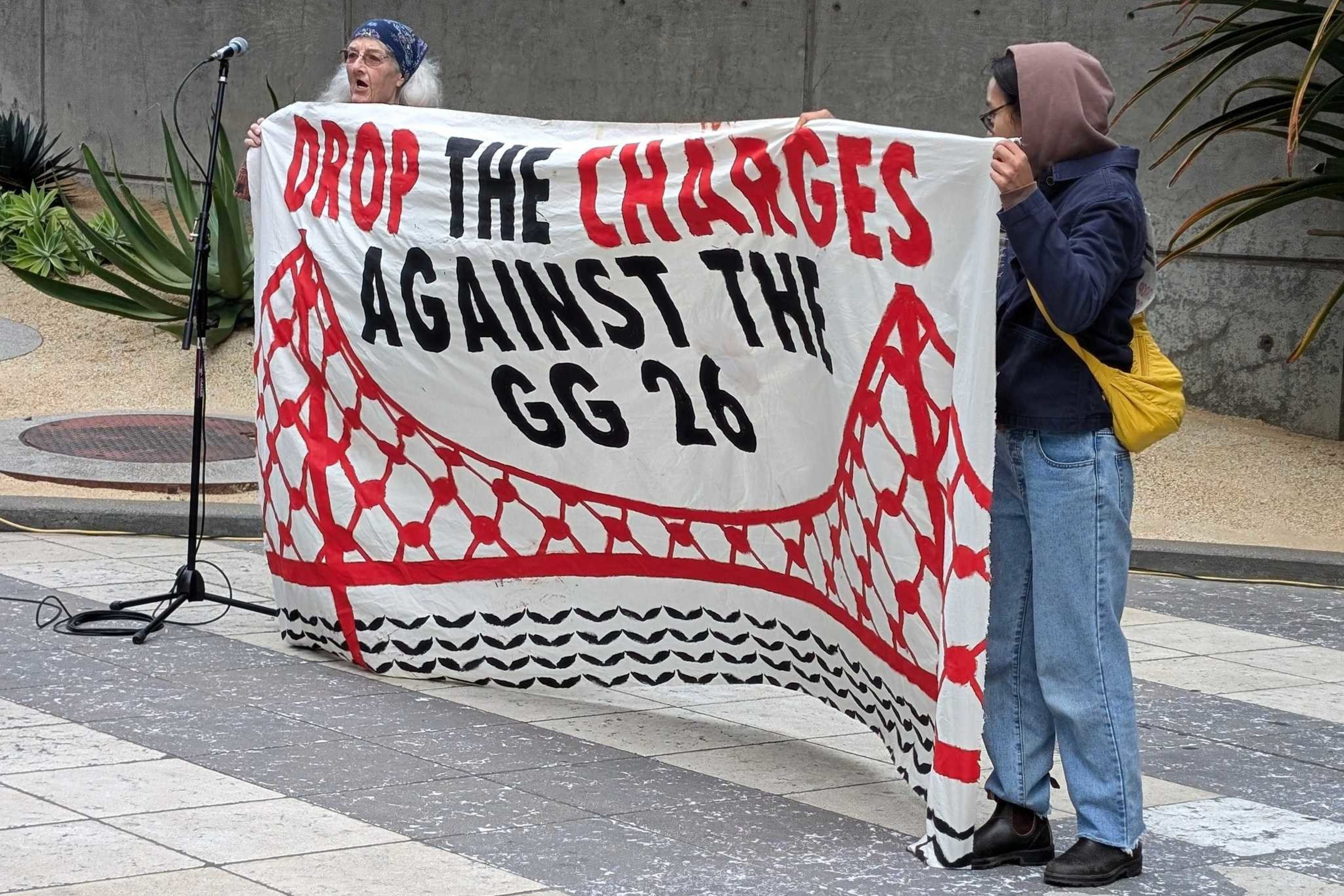 Two people hold a banner that says "drop the charges against the GG 26," referring to the 26 people facing charges from a demonstration that blocked traffic on the Golden Gate Bridge in April.