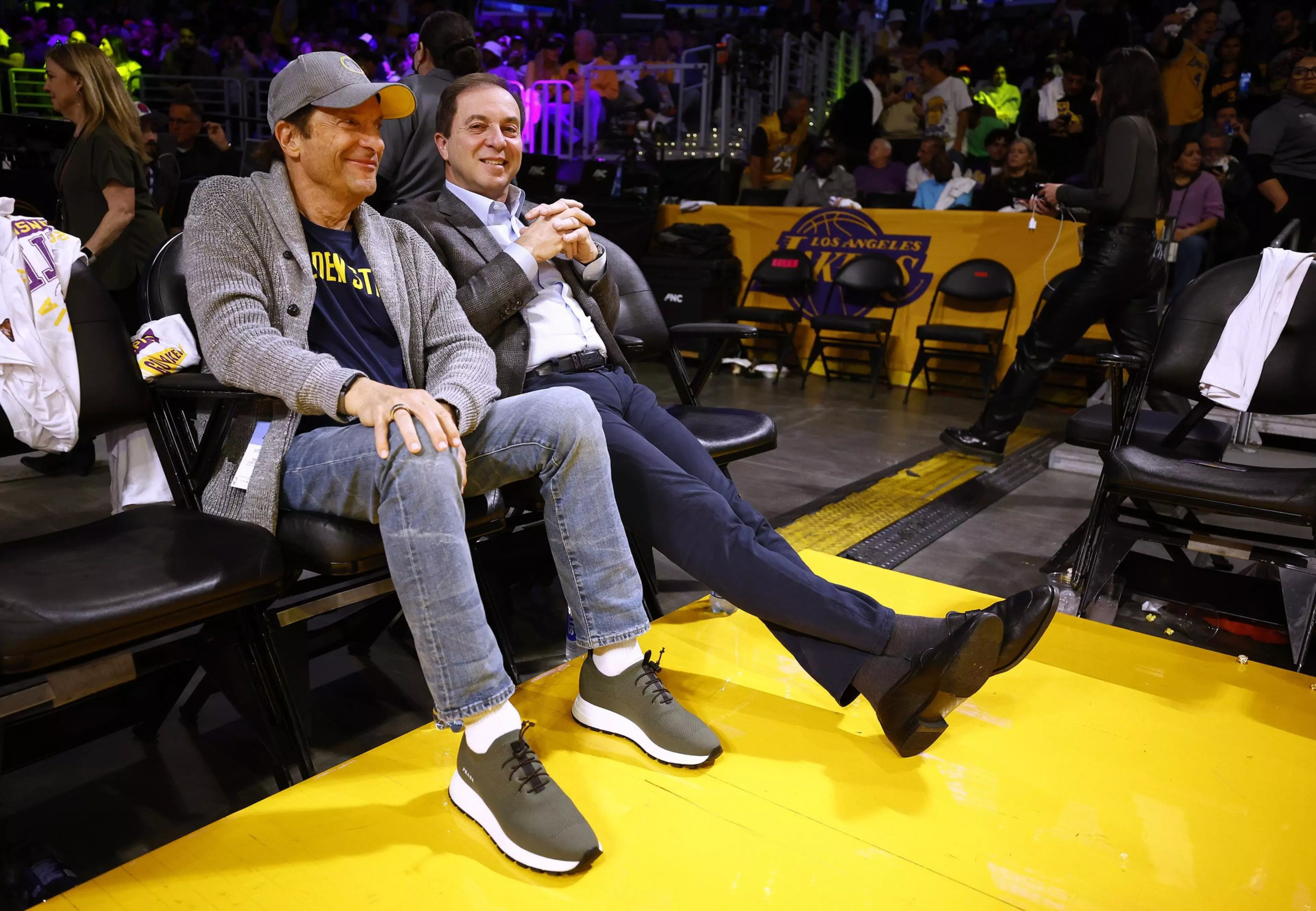 Two men sit courtside at a basketball game, smiling and relaxed, with one wearing casual clothes and sneakers and the other in a suit and dress shoes.