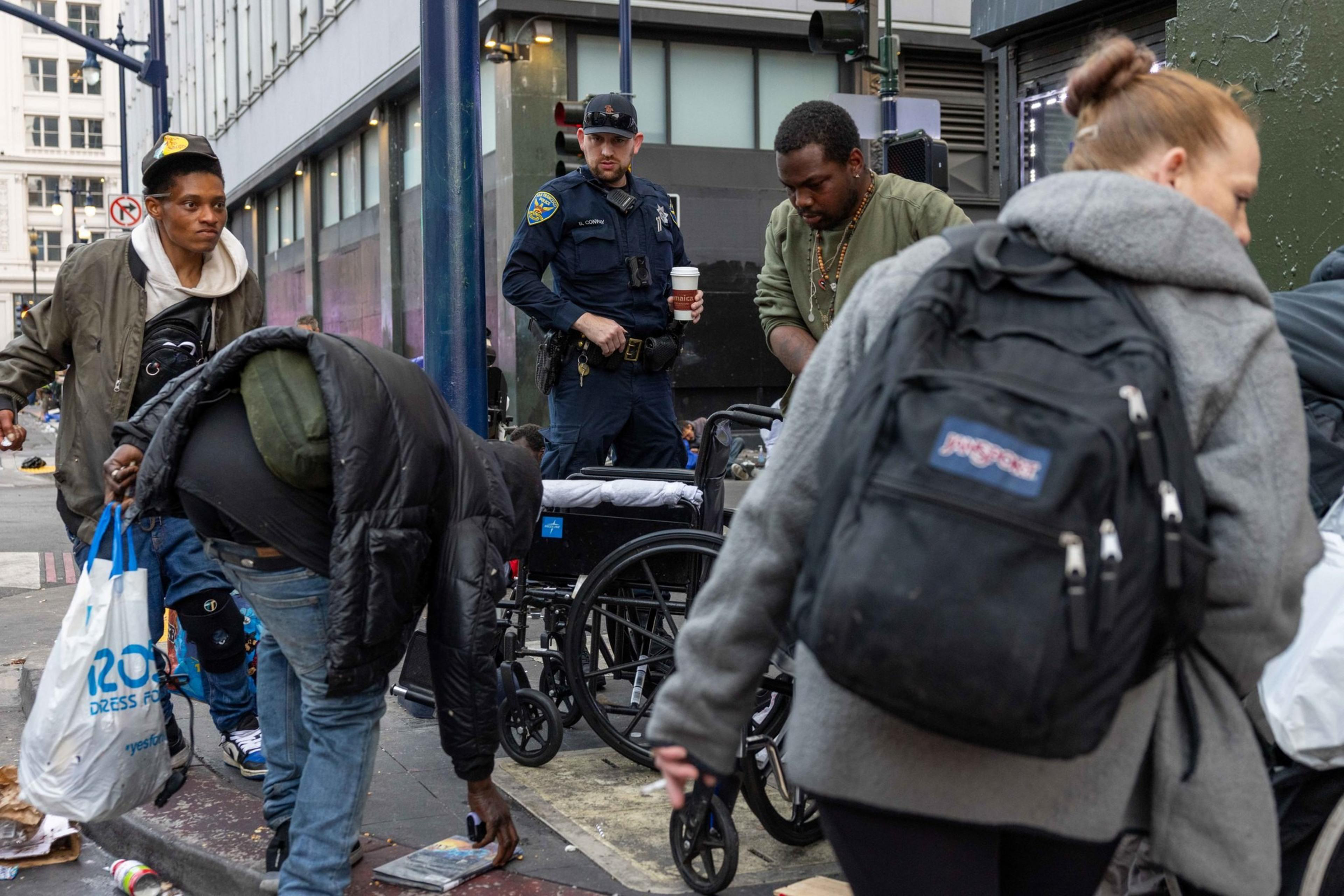 A city sidewalk with people, including a police officer holding a coffee, a man bending to pick up an item, others with backpacks, and a wheelchair nearby.