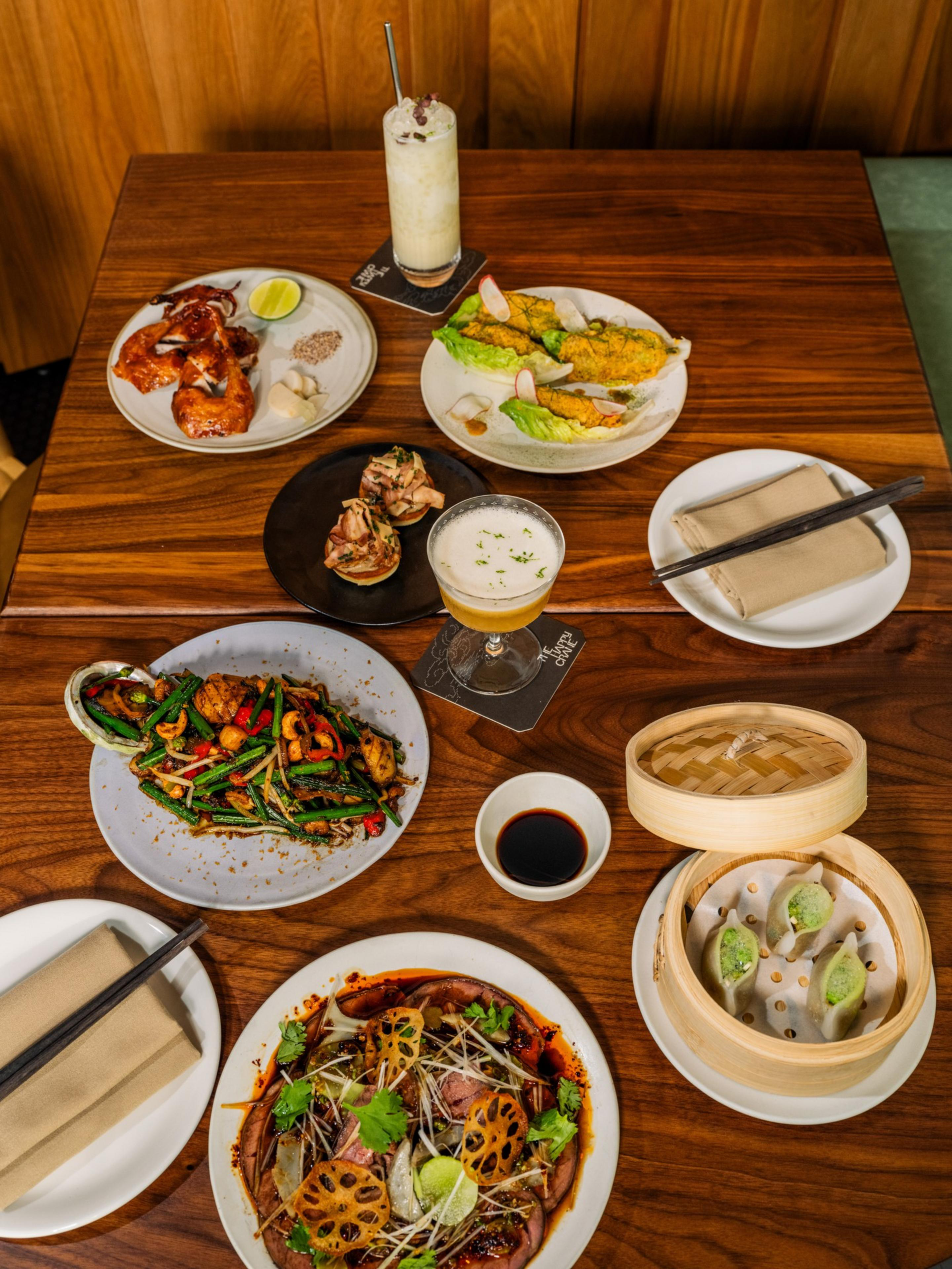 Grilled chicken, stuffed lettuce wraps, sautéed vegetables, dumplings in bamboo steamer, spicy meat dish with lotus root, two cocktails, soy sauce, and chopsticks on a wooden table.
