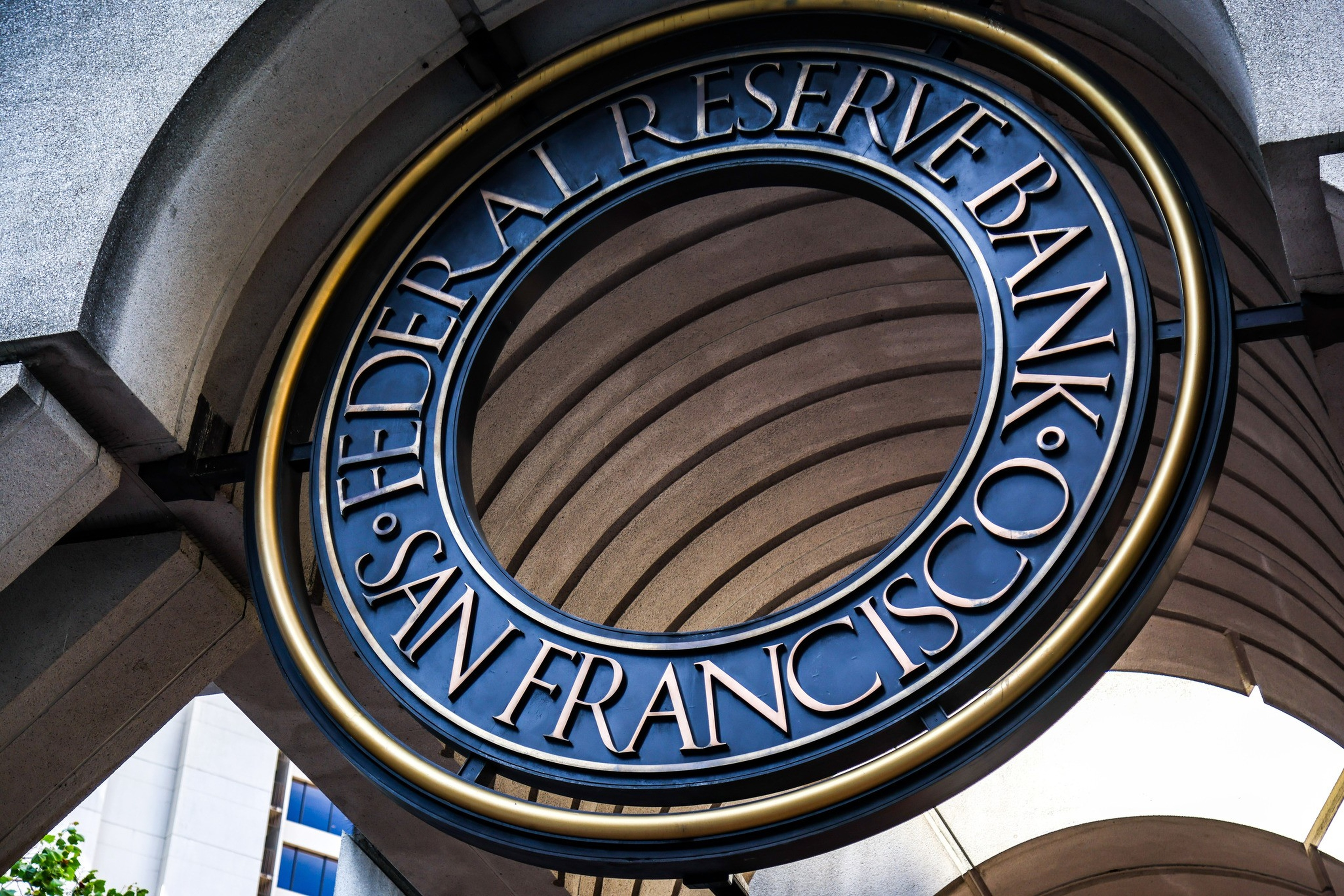 A circular metallic sign reads "Federal Reserve Bank San Francisco," hanging under a stone archway with curved lines.