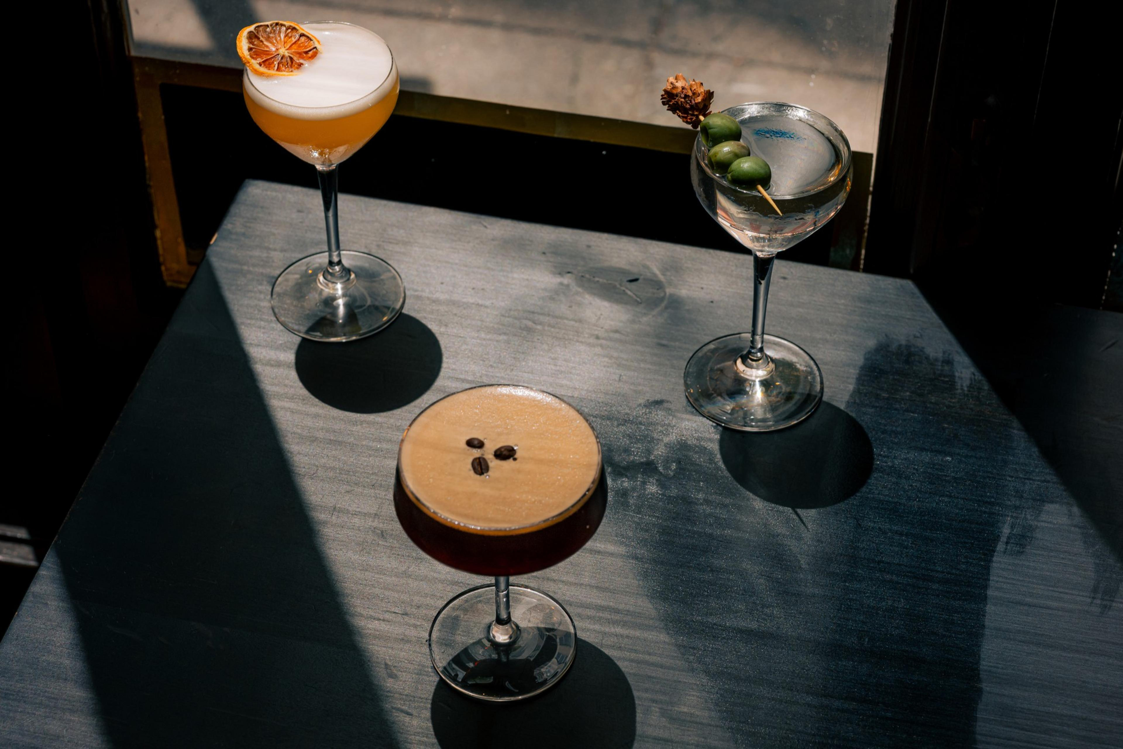Three cocktails on a table: an orange-topped one, an espresso martini with coffee beans, and a clear one with olives. Sunlit with distinct shadows.