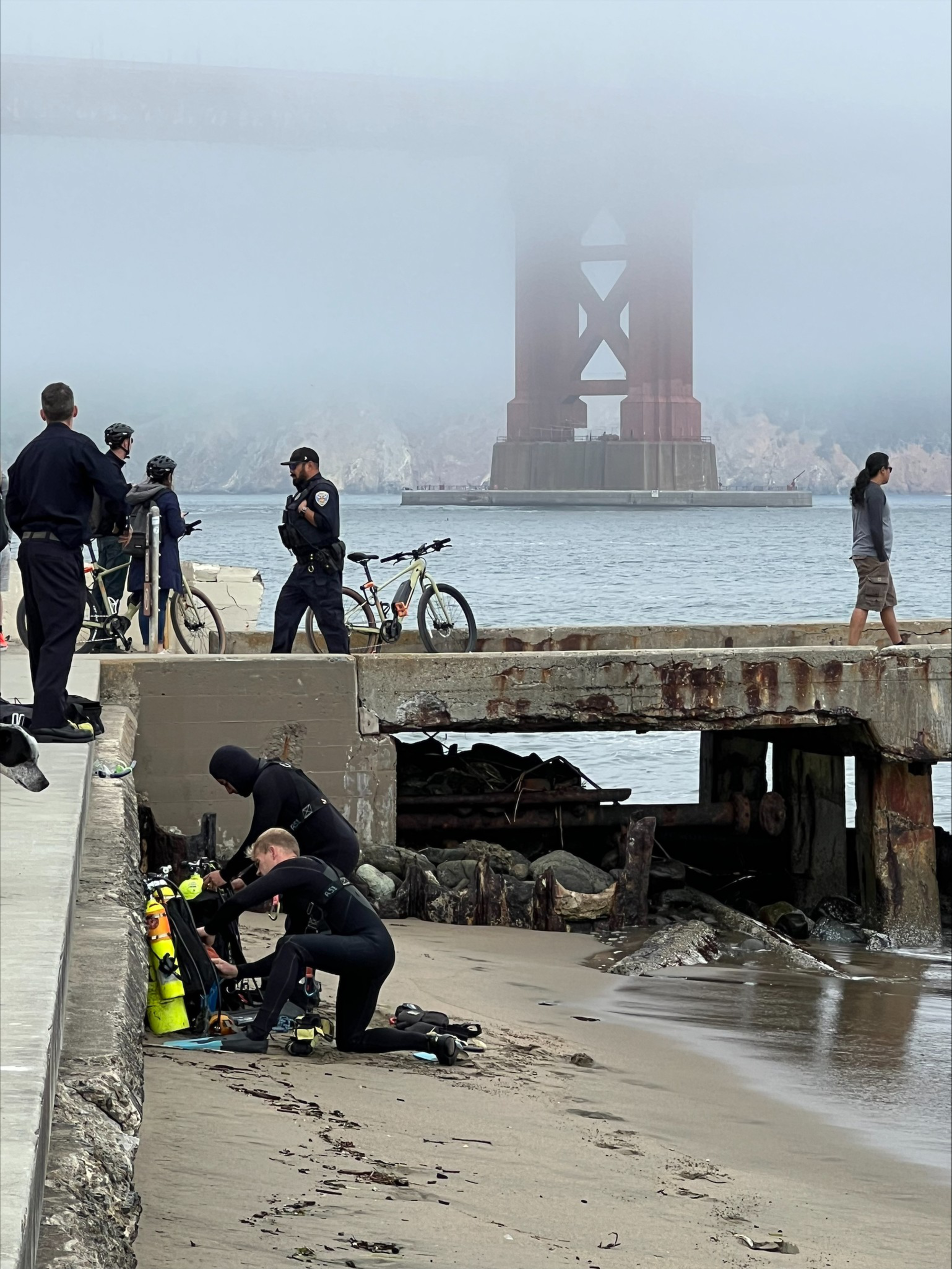 Two divers prepare gear on a sandy shore beneath a rusty pier, while police and cyclists stand nearby with the fog-covered Golden Gate Bridge in the background.