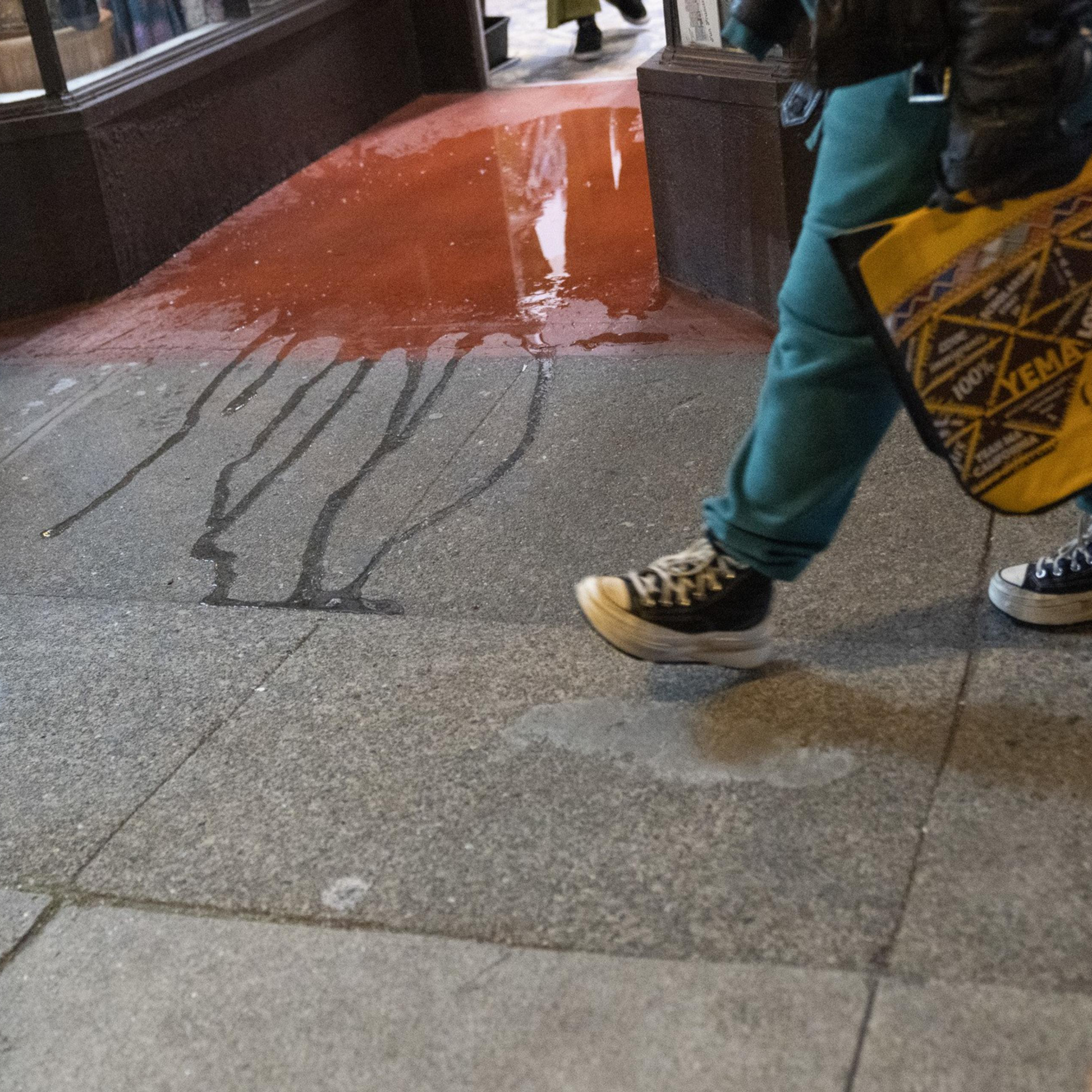 A person walks past a storefront with reddish liquid spilled on the sidewalk. They carry a yellow bag and wear blue pants and black sneakers.