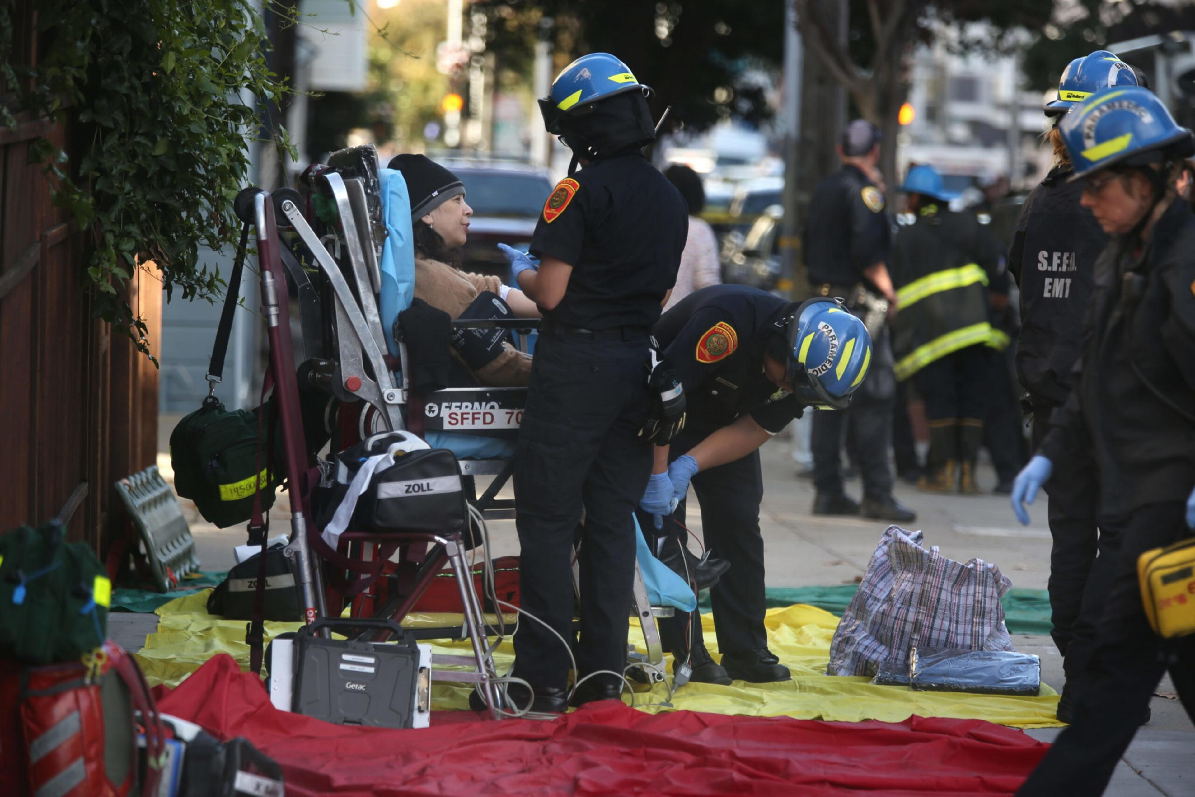 Emergency medical personnel wearing helmets and gloves assist a seated elderly woman on a sidewalk with medical equipment and bags nearby.