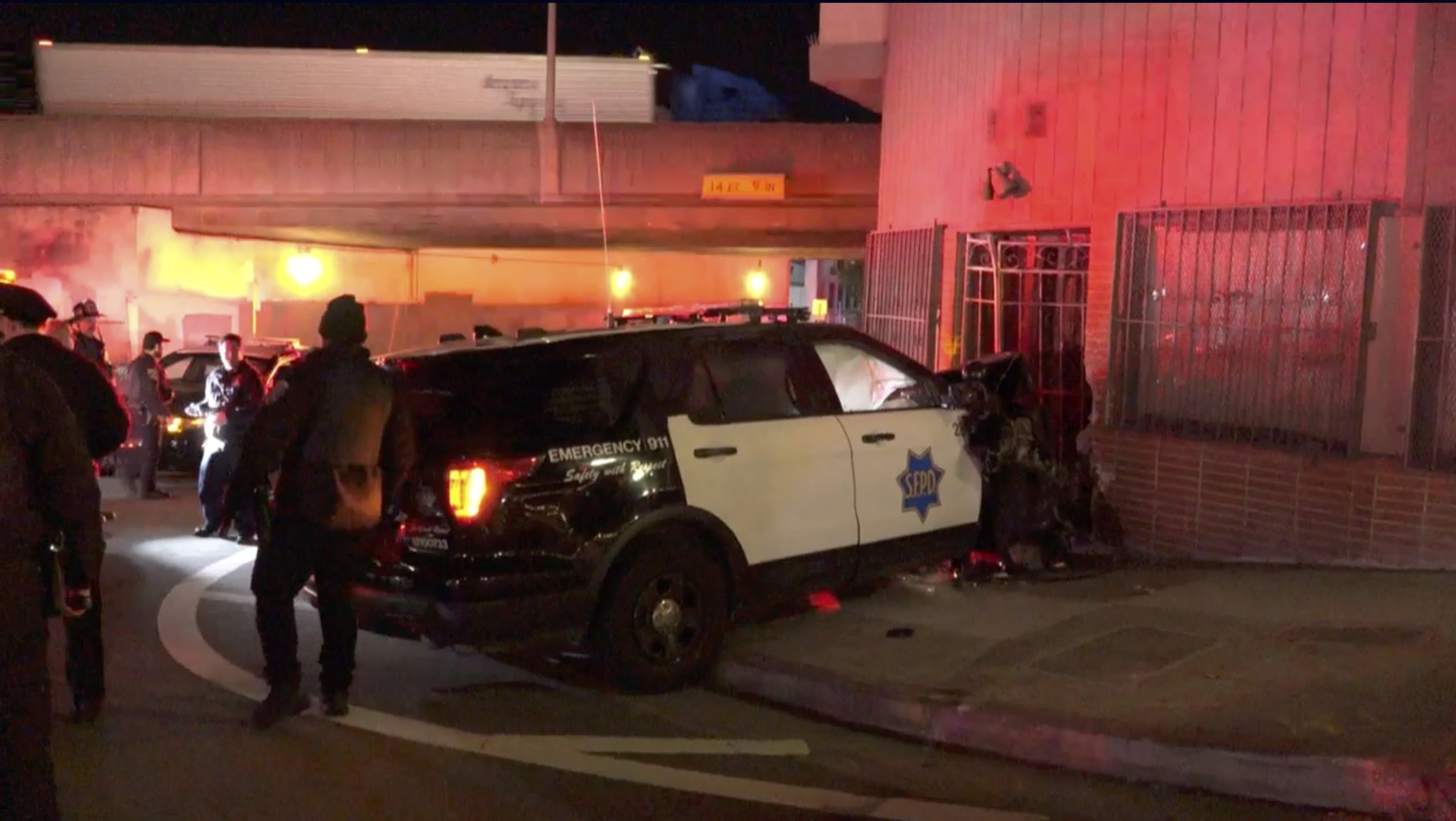 A police car has crashed into a building at night. Several officers are present, with red emergency lights illuminating the scene.