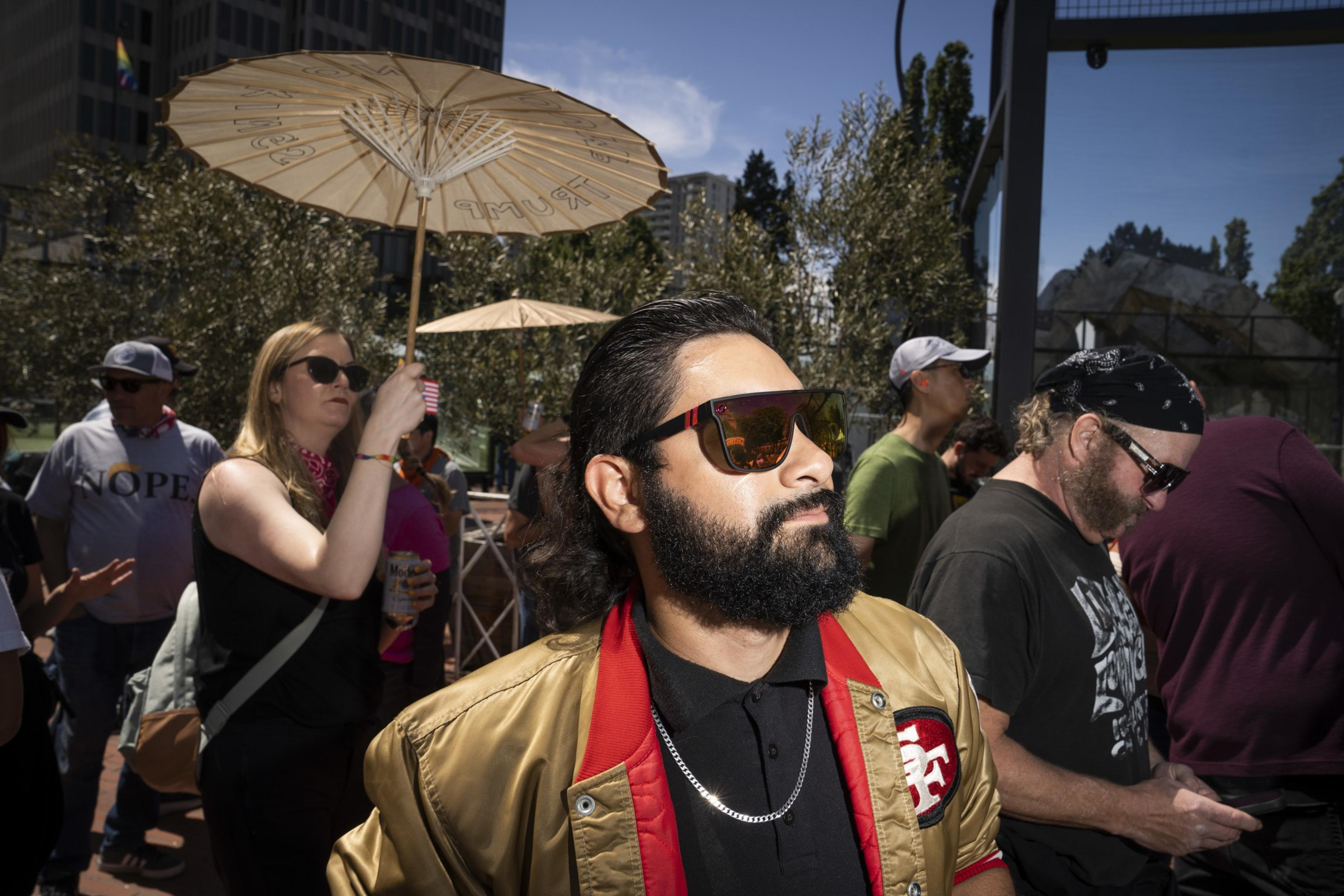 A bearded man in sunglasses and a gold jacket is in the foreground, with people around him holding umbrellas and drinks, under a clear blue sky.