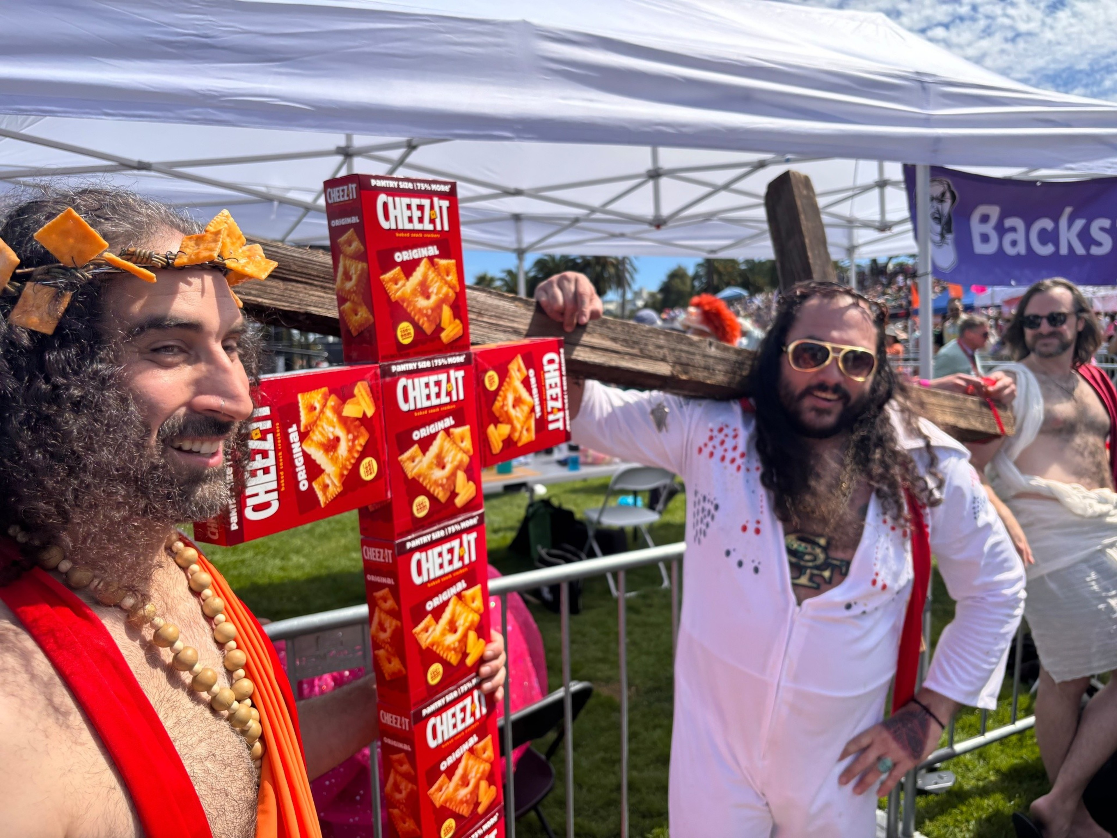 Two men hold a wooden cross adorned with Cheez-It boxes, one wearing a cheese cracker crown and beads, the other in a white outfit with sunglasses.
