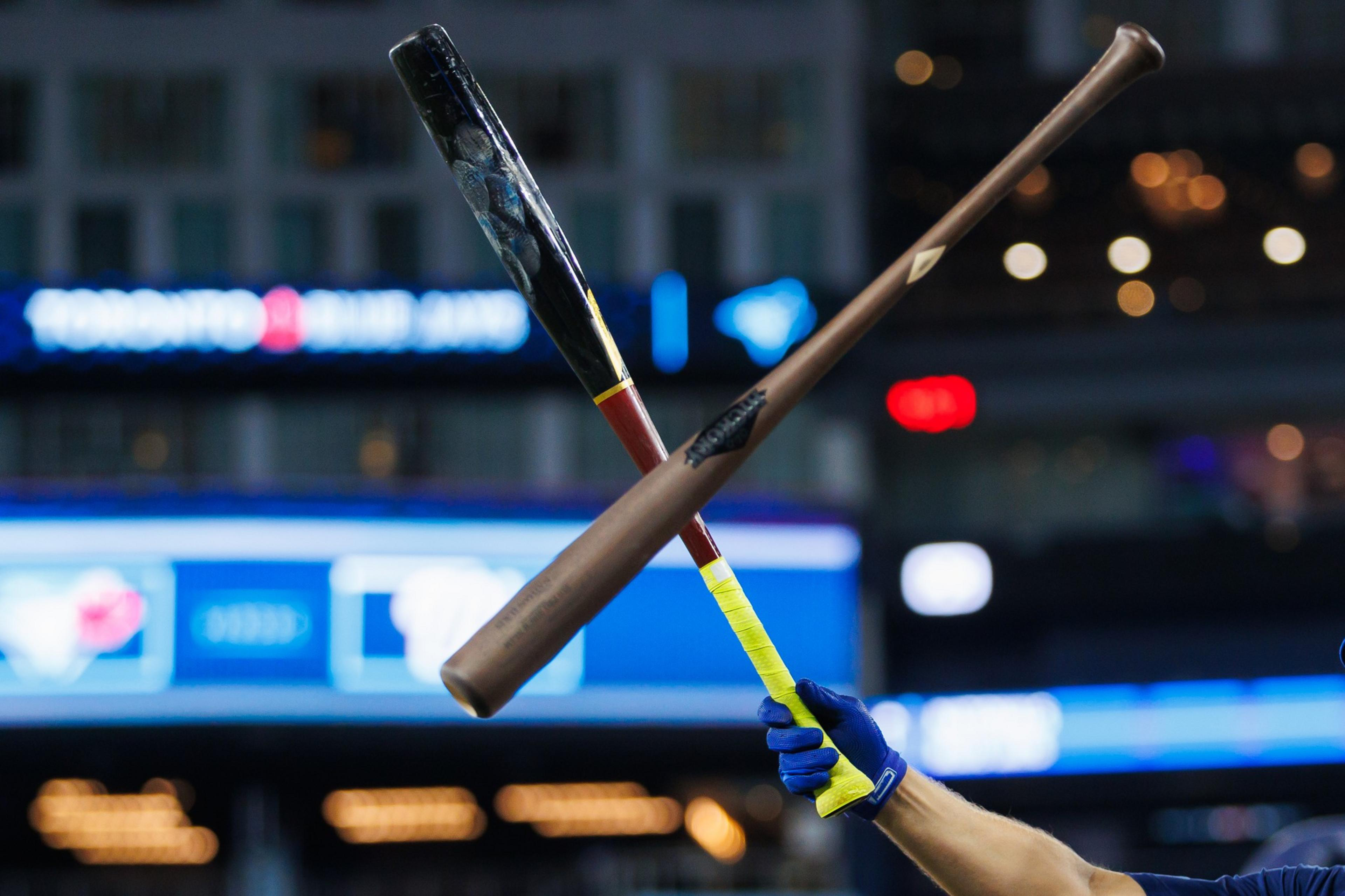 A person wearing a blue glove holds two crossed baseball bats with yellow grips, set against a blurred stadium background displaying &quot;TORONTO BLUE JAYS.&quot;