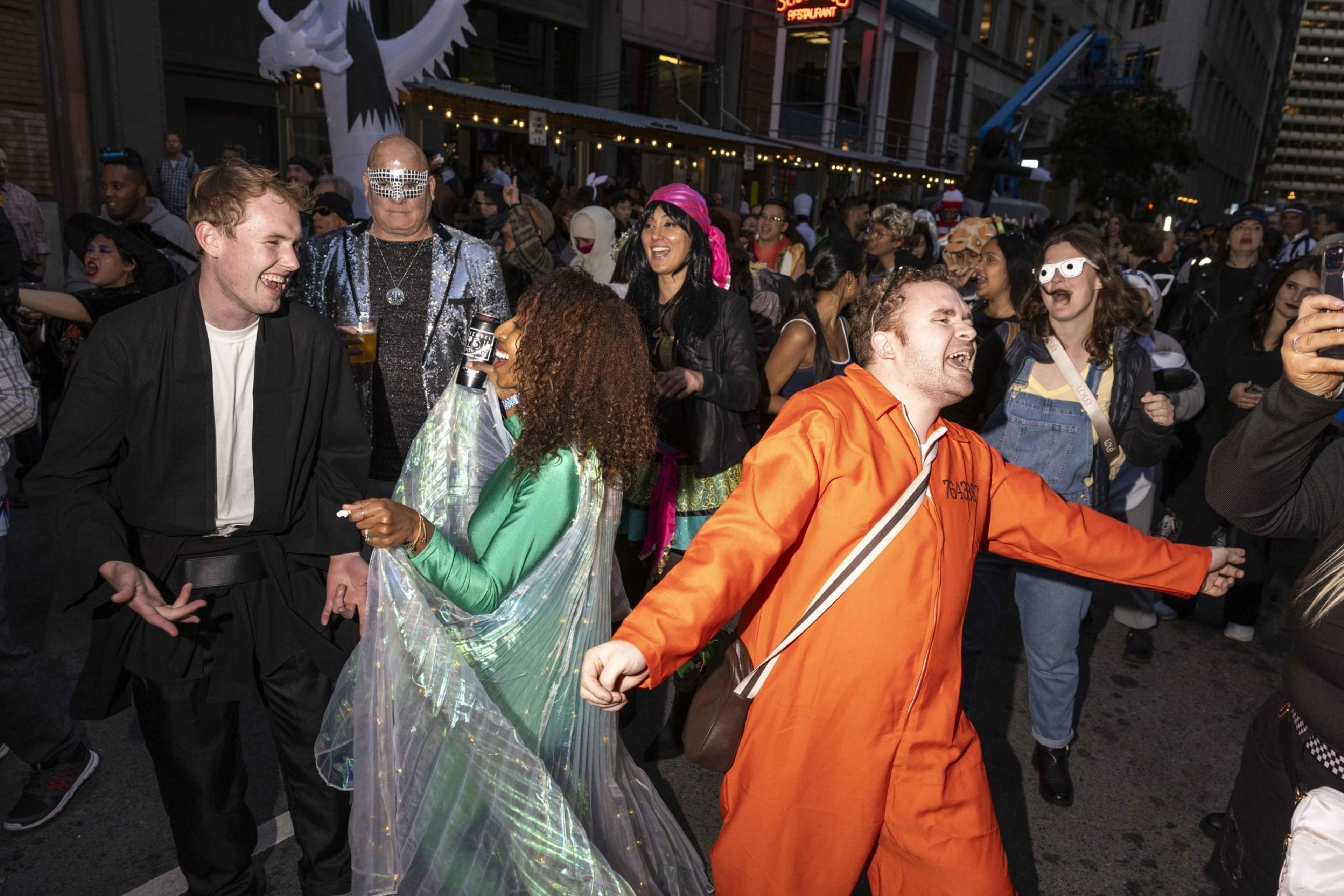 A lively street party with people in elaborate costumes, including a man in an orange jumpsuit and others wearing metallic and colorful outfits, all enjoying the event.