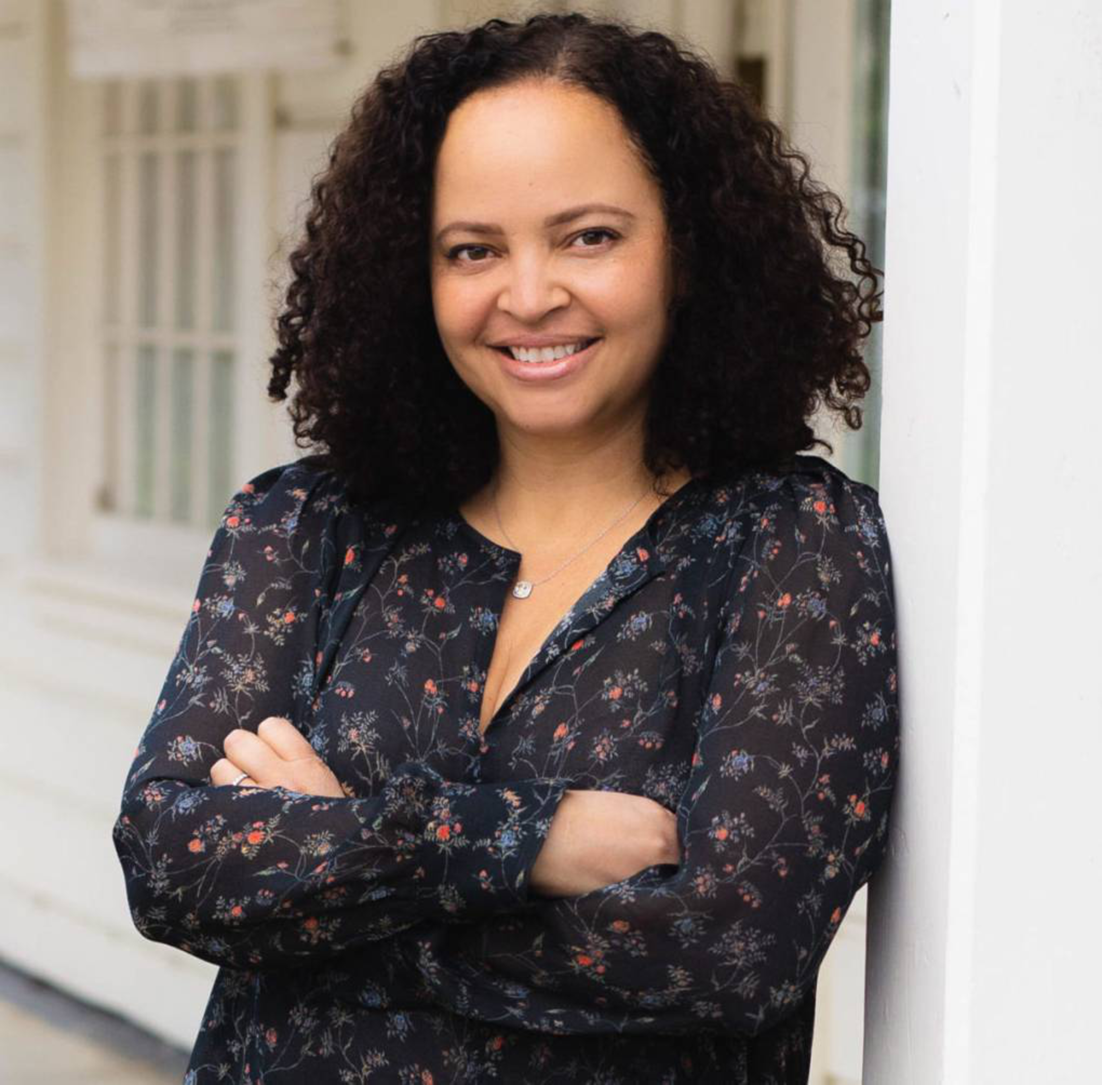 A woman with curly hair smiles, standing with arms crossed. She wears a dark, floral-patterned blouse and leans against a white structure, with windows behind her.
