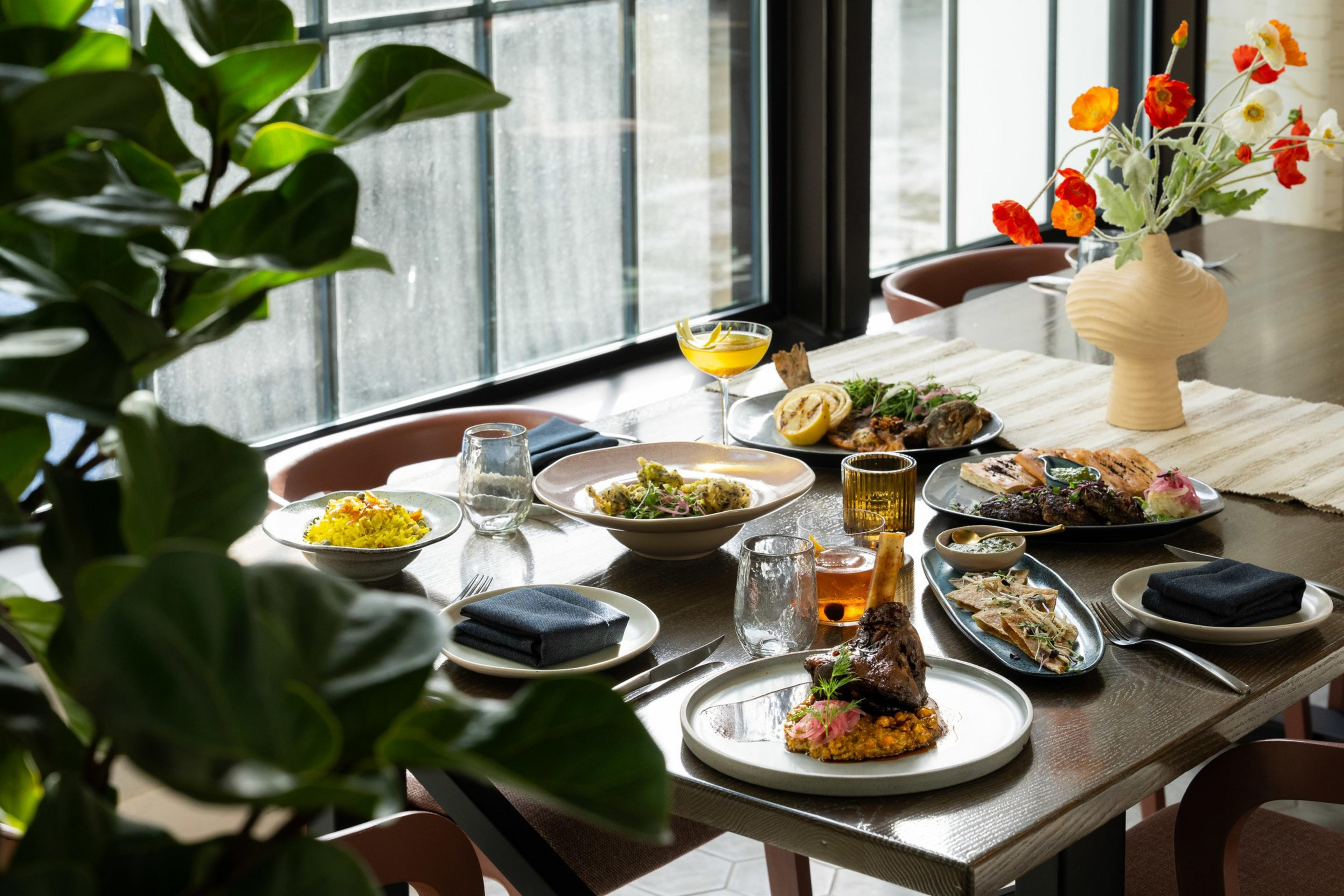 The image shows a dining table set with various dishes, glasses, and a vase with red and white flowers. The scene is lit by natural light from large windows.