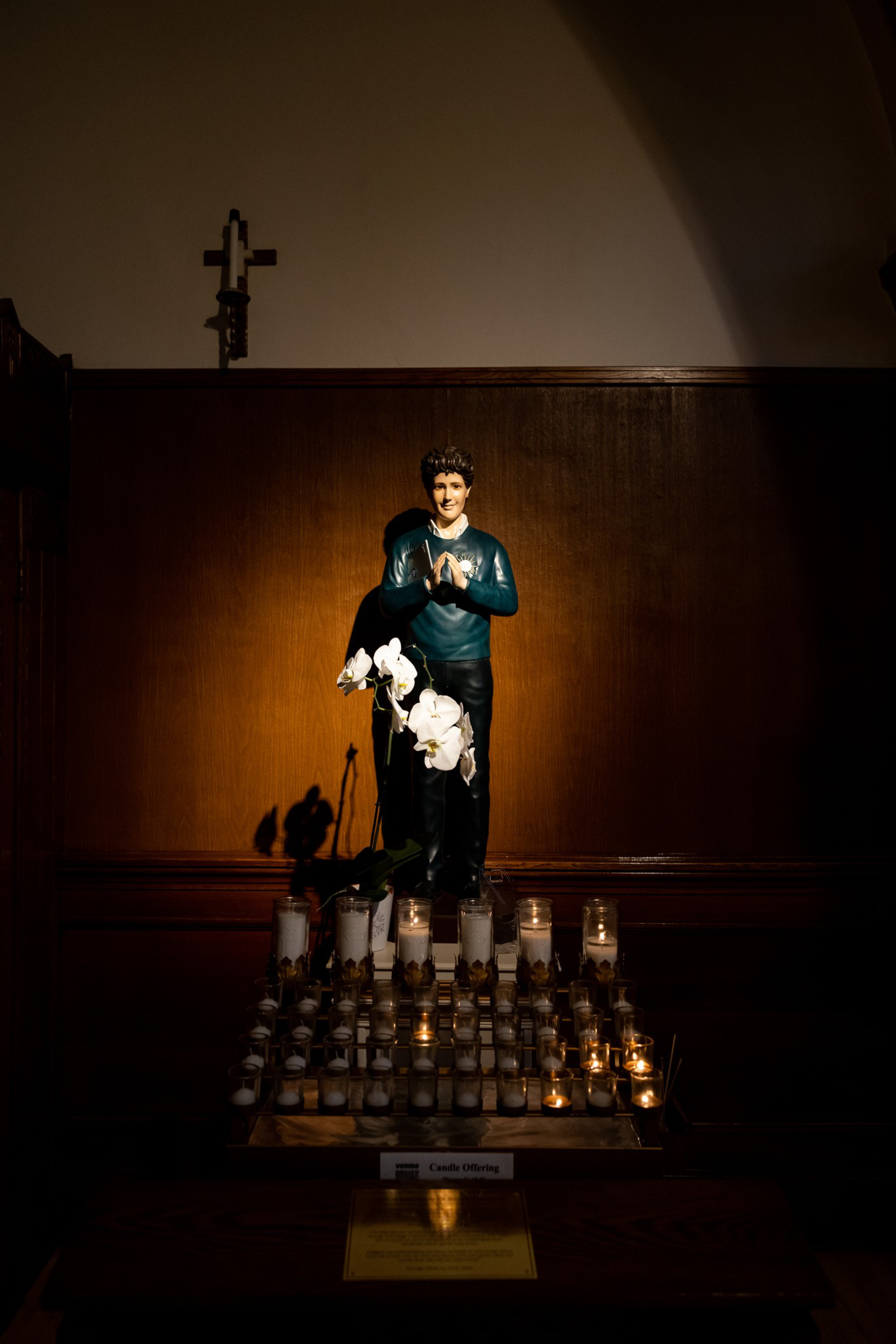 A statue of a person with hands clasped in prayer stands behind white flowers and numerous lit and unlit candles on a wooden altar.
