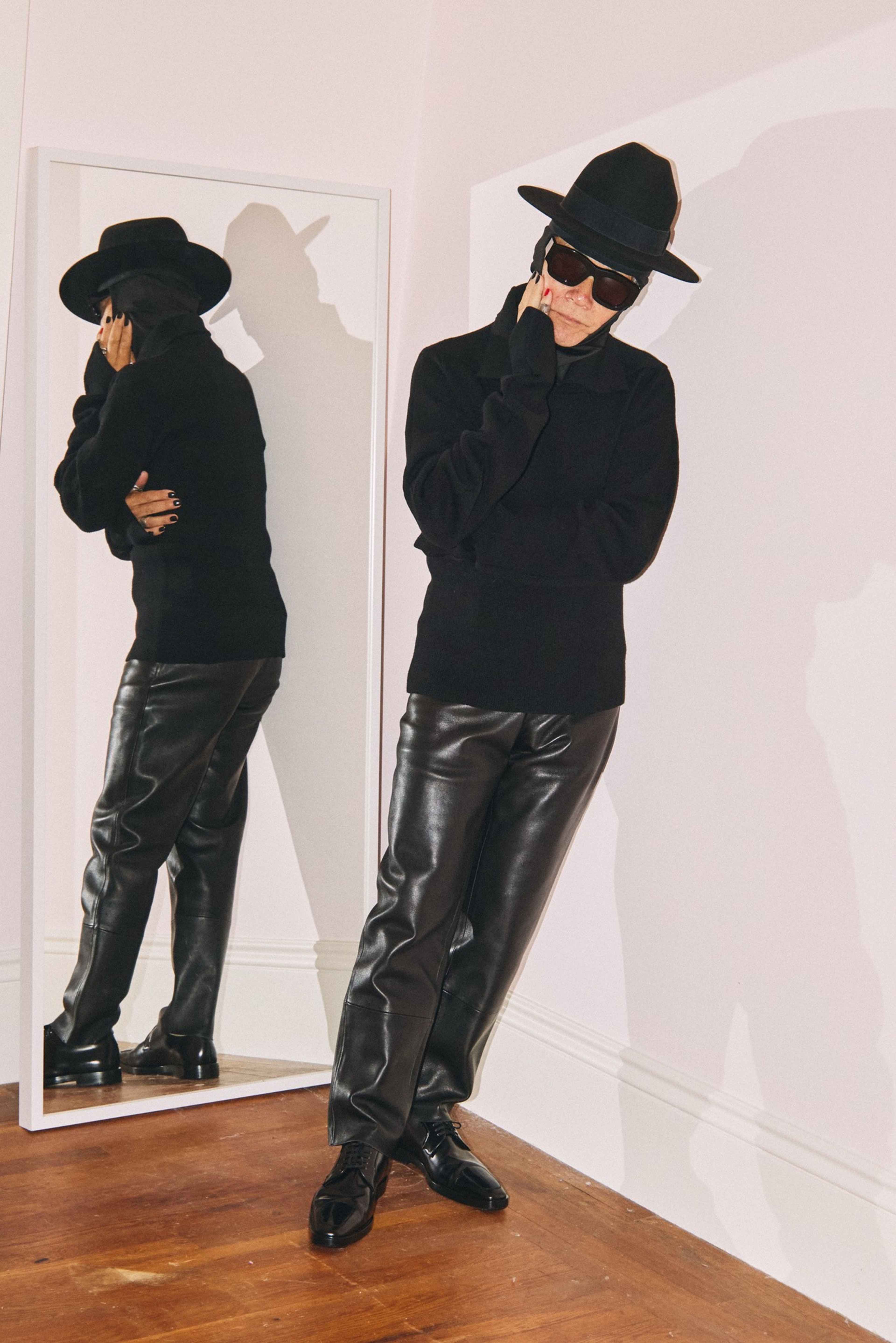 A person dressed in black leather pants, a dark sweater, hat, and sunglasses poses with their hand on their chin, standing in front of a mirror.