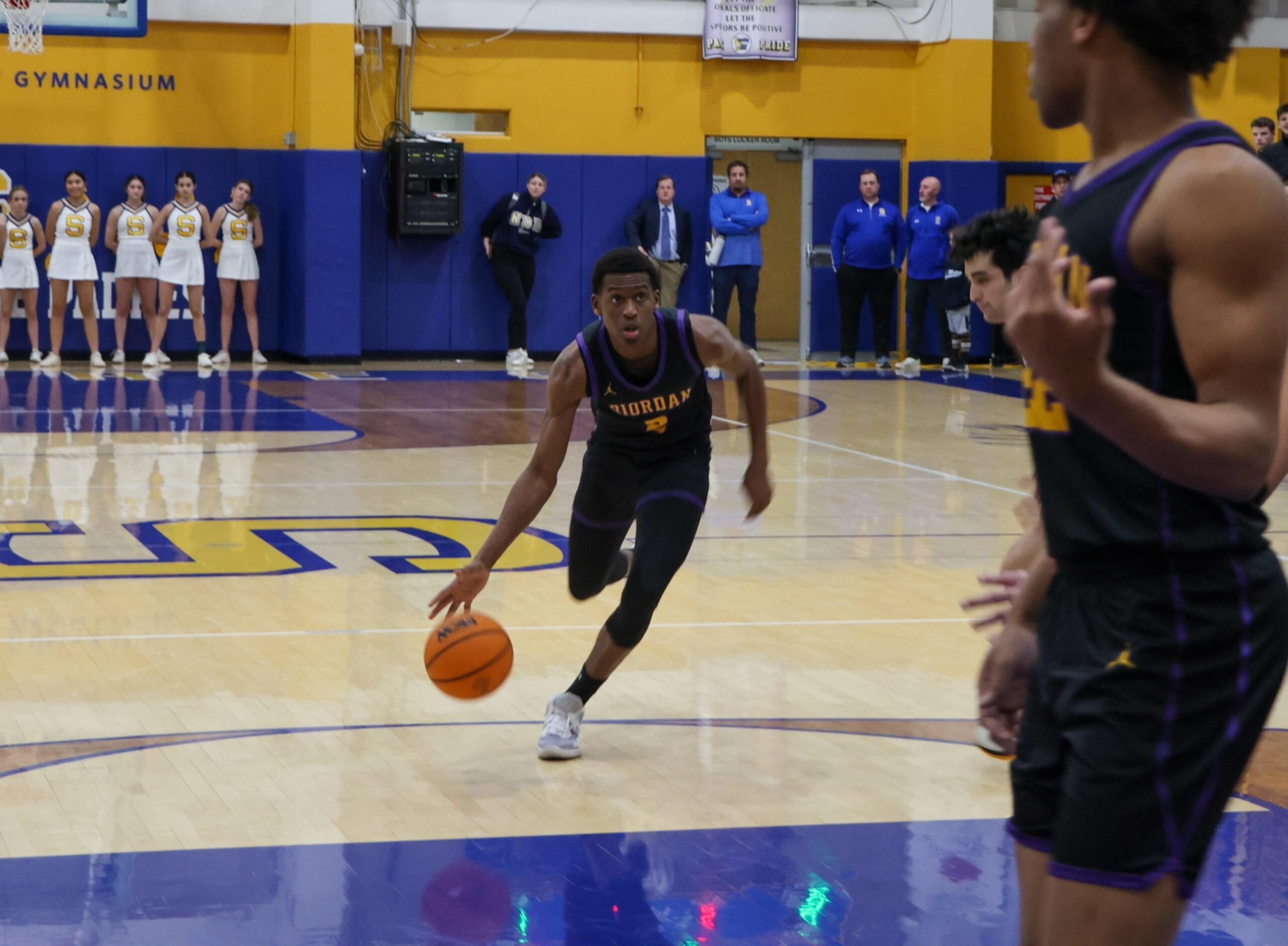 A basketball player in a black "Jordan" uniform dribbles the ball toward the basket, with teammates, cheerleaders, and coaches watching from the sidelines.