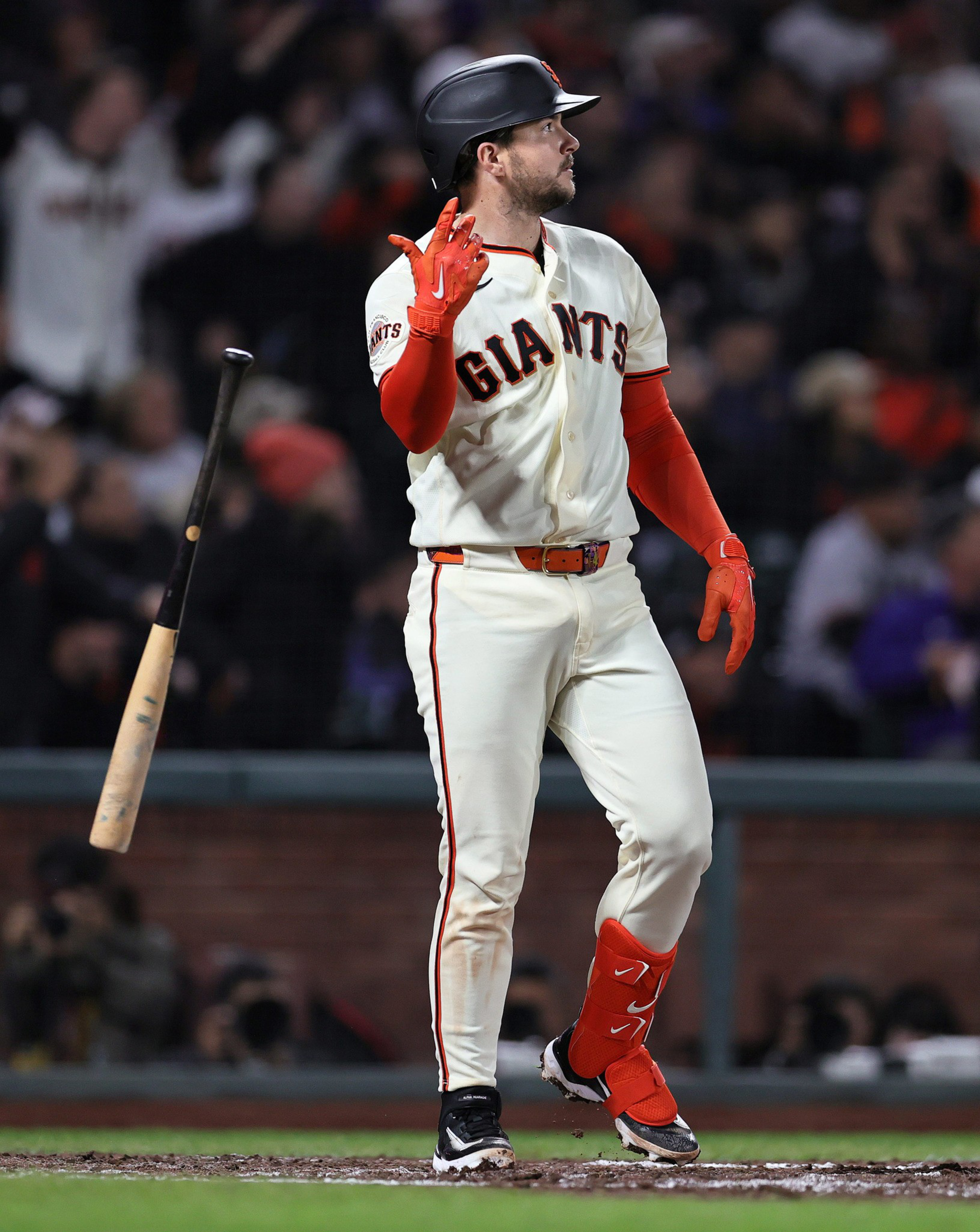 A baseball player in a white Giants uniform with red gloves and accessories is tossing a bat while standing at home plate.