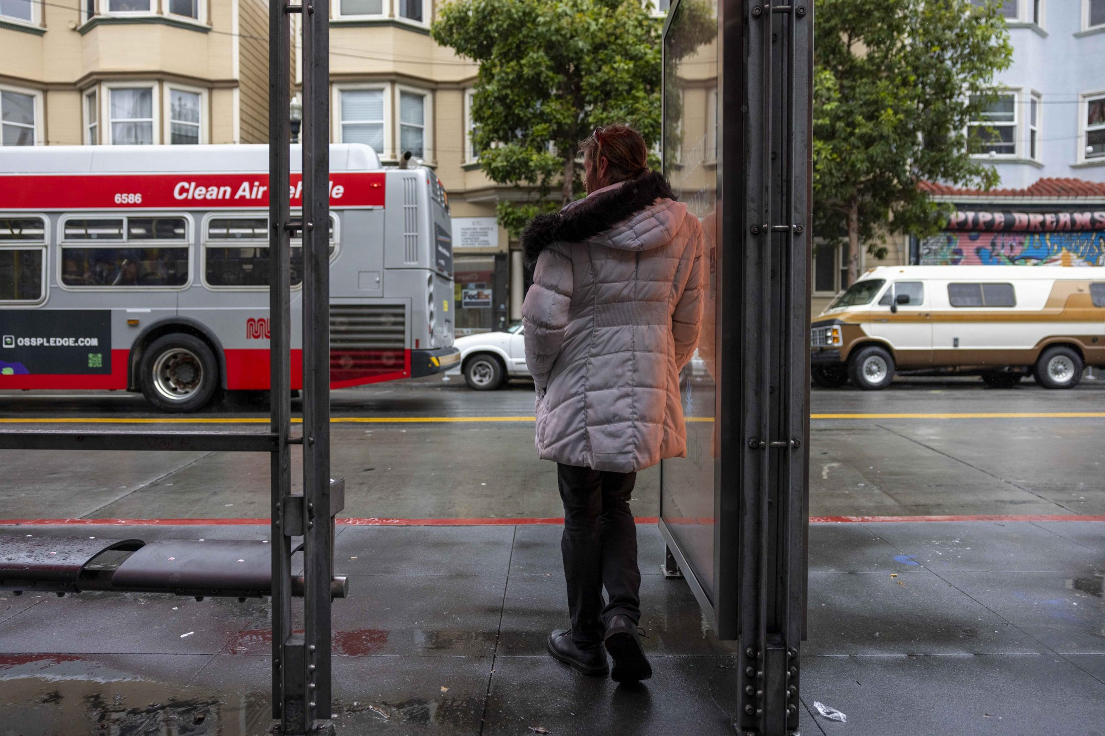 A person in a white coat stands at a bus stop, with a red and white bus passing by on a wet street. A tree and buildings are visible in the background.