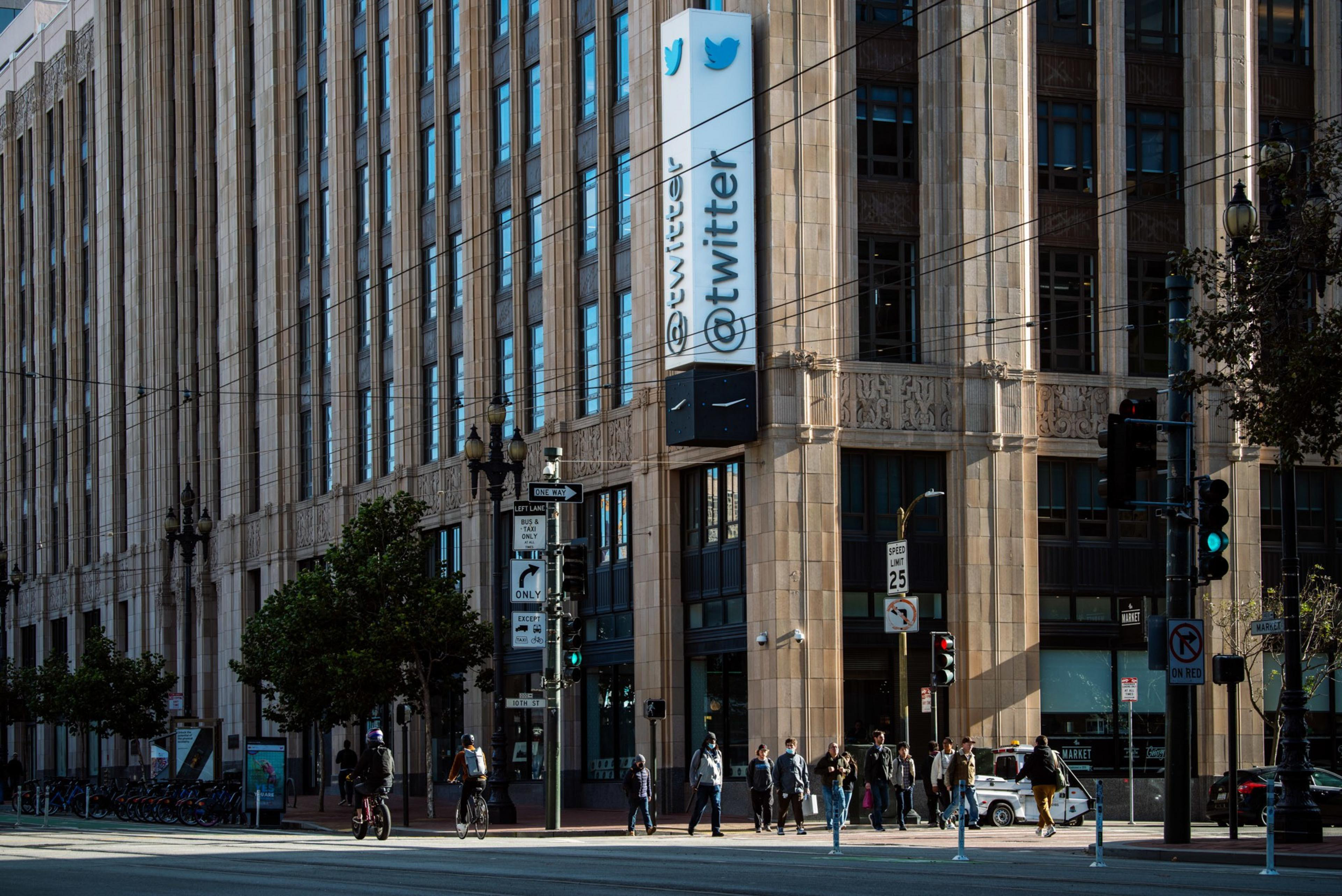 People walk and bike past a large building featuring a vertical sign with the Twitter logo and handle. The scene includes trees and street signs.