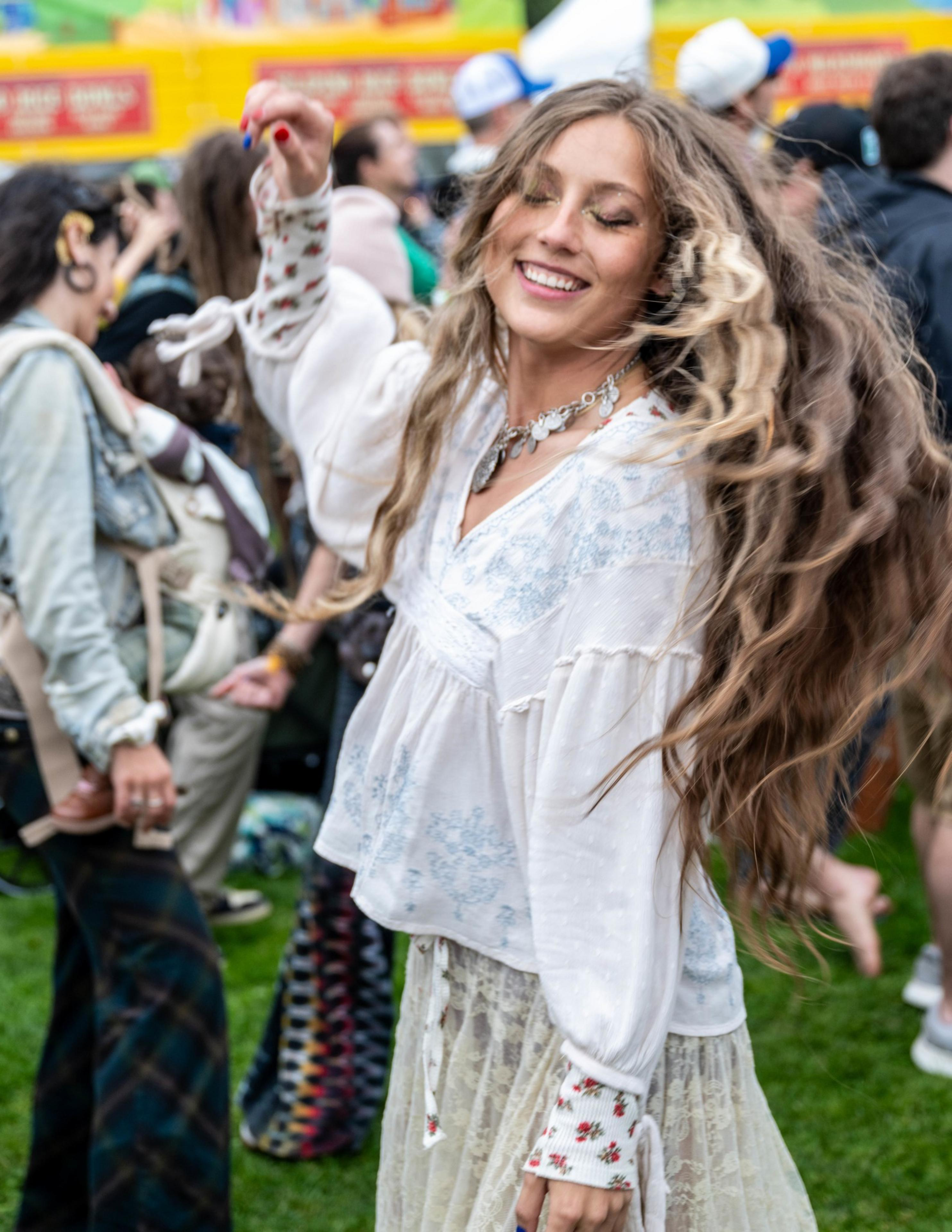 A smiling woman with long wavy hair twirls, wearing a white blouse and floral skirt, surrounded by people at an outdoor event.