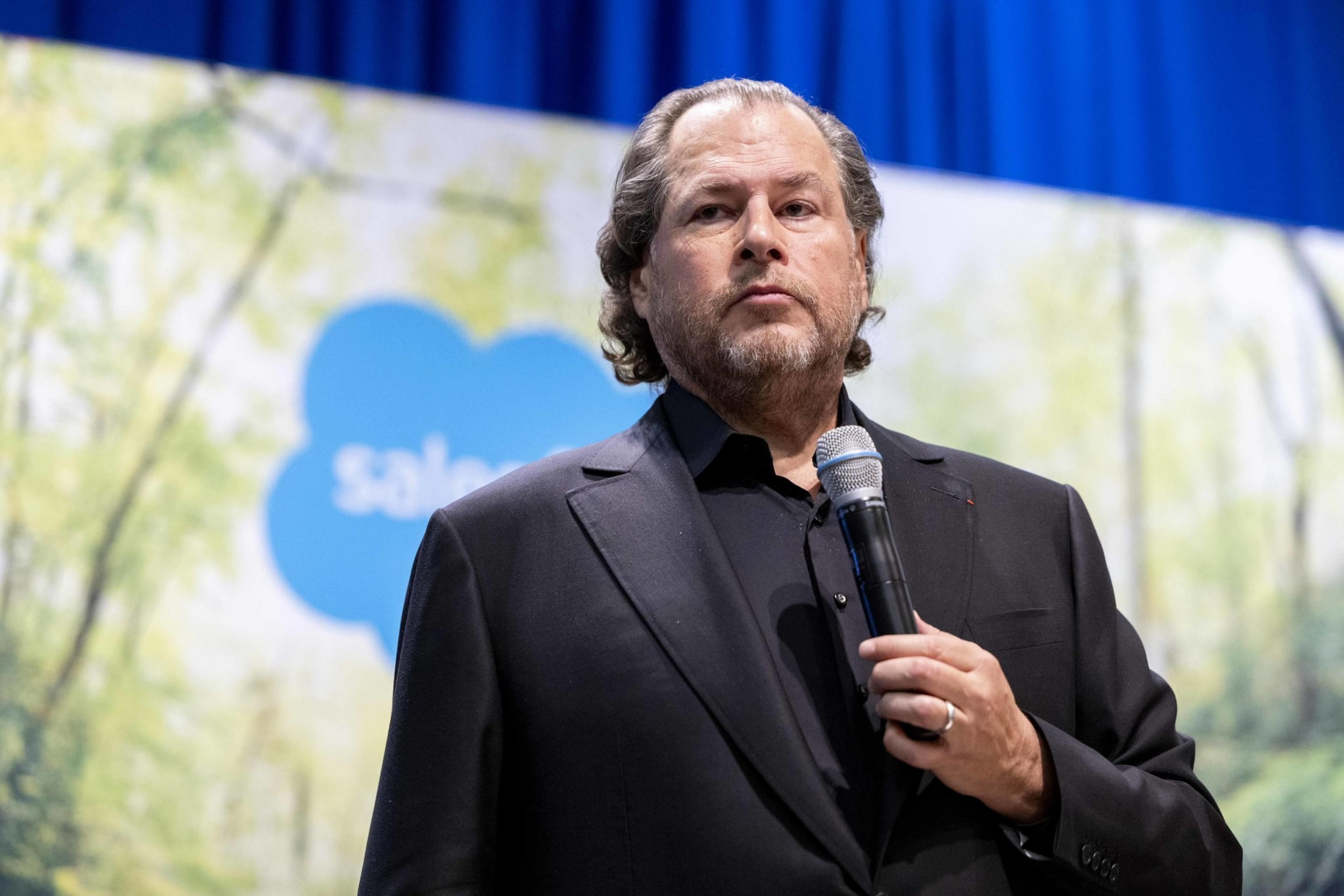 A person in a black suit holds a microphone, standing in front of a backdrop with greenery and a blue logo.