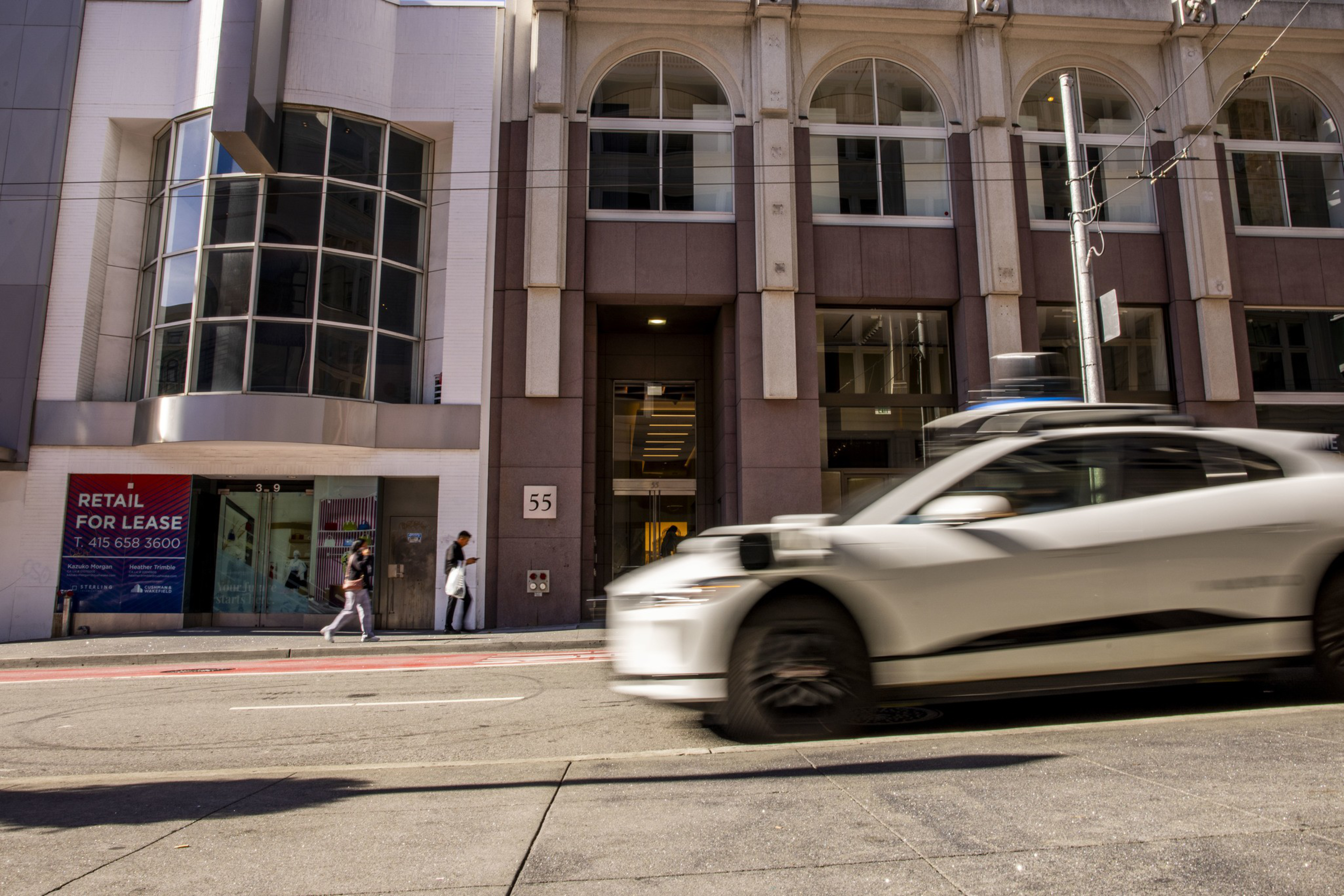 A white autonomous car is driving past a building with large arched windows, a “Retail for Lease” sign, and two people walking on the sidewalk.