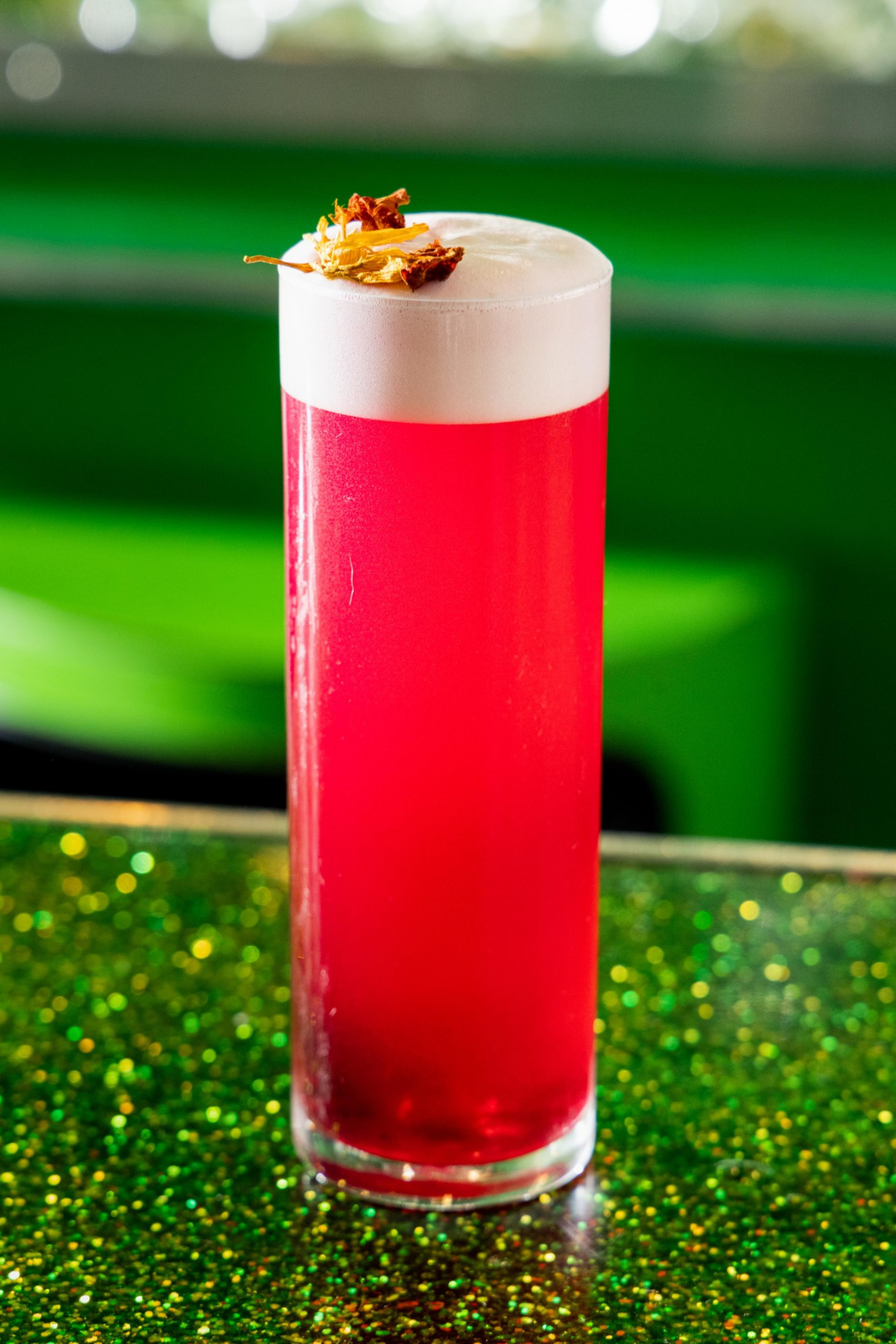 A tall glass holds a bright pink frothy drink, topped with a small garnish of dried flowers, placed on a sparkly green surface.
