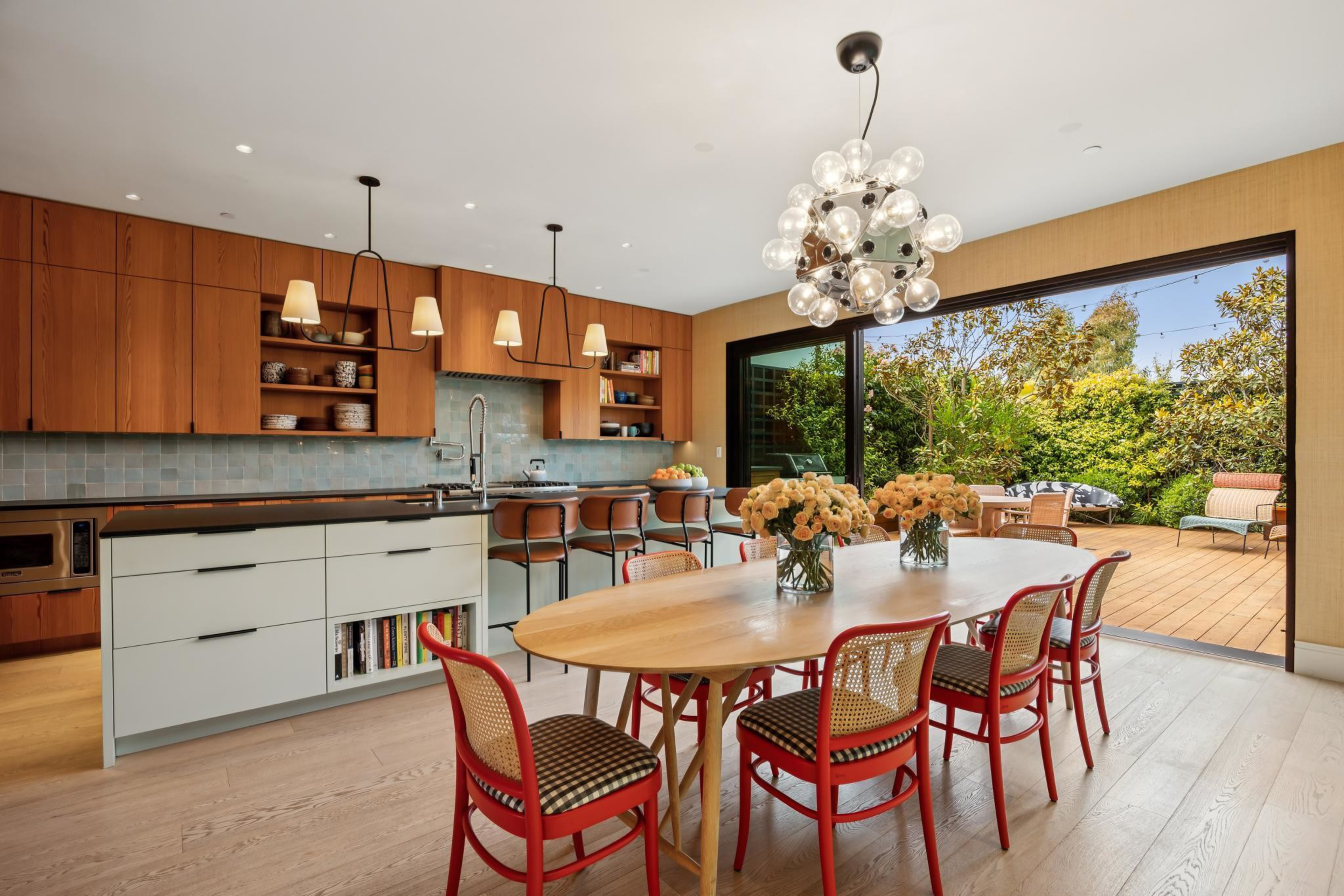 A modern kitchen and dining area with a wooden table, red chairs, pendant lights, a large sliding glass door opening to a deck with outdoor seating and greenery.