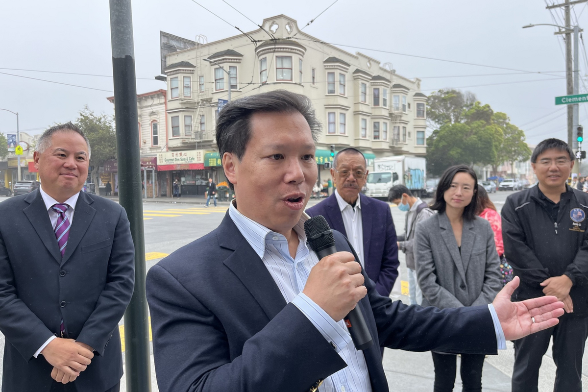 A man speaks into a microphone on a city street, with four onlookers behind him, building and foggy sky in the background.
