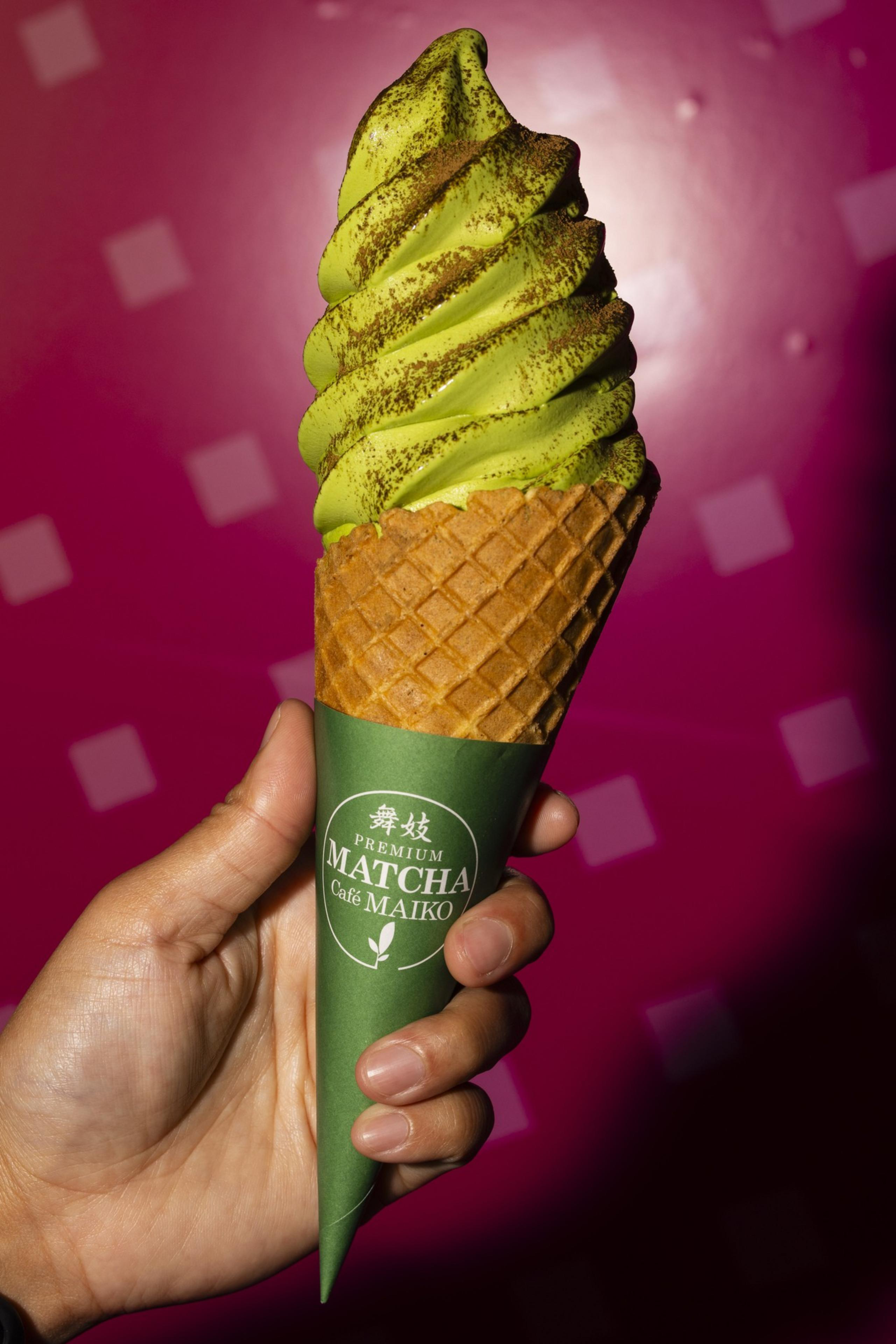 A hand holds a waffle cone filled with green matcha soft-serve ice cream, topped with a sprinkle of powder, against a pink background.
