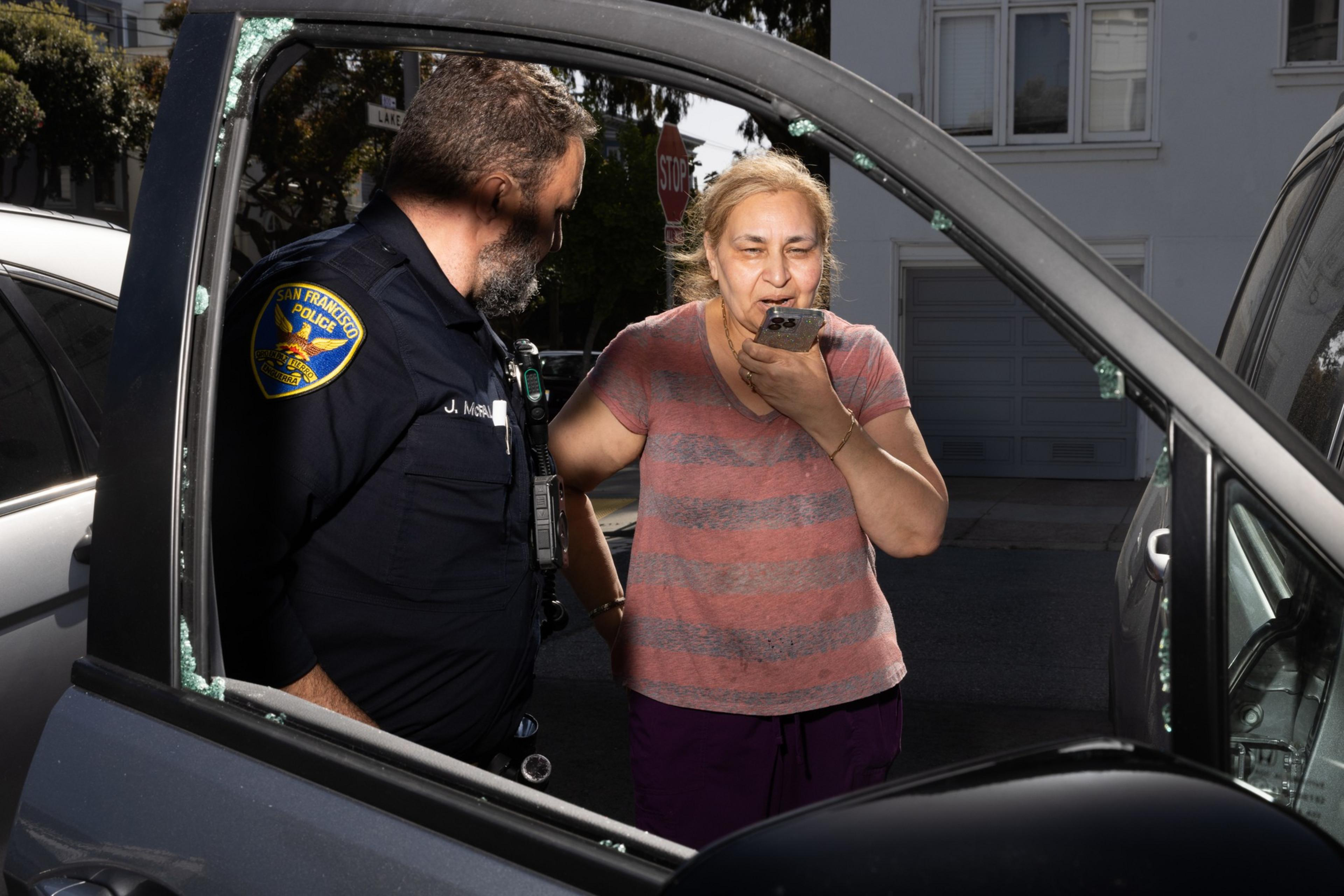 A woman points her smartphone at a police officer by a car with a shattered window, likely recording him. The officer looks at her phone.