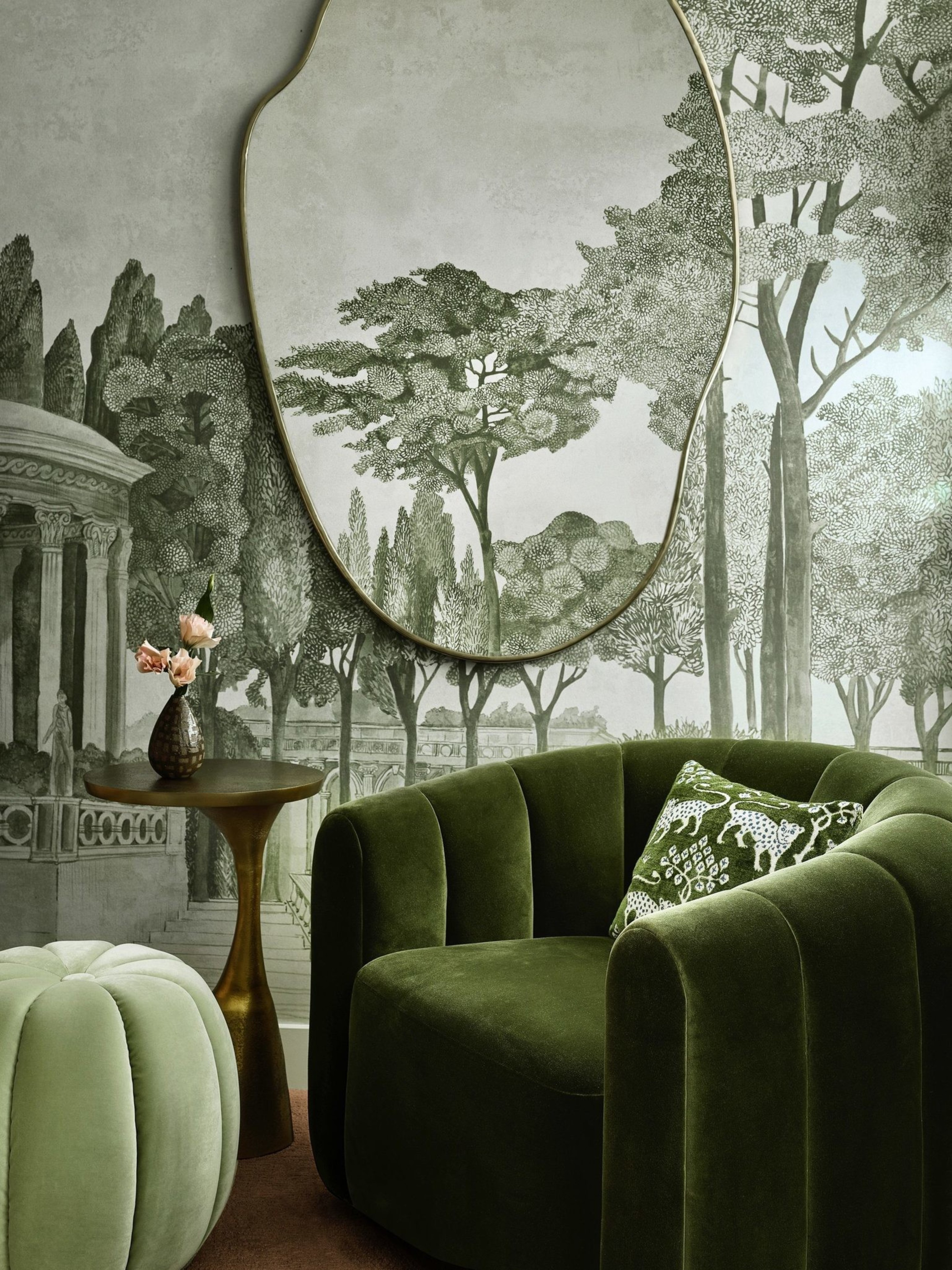 A room with a green velvet armchair, a matching pouffe, a gold side table with flowers, and a large mirror reflecting a detailed mural of trees.