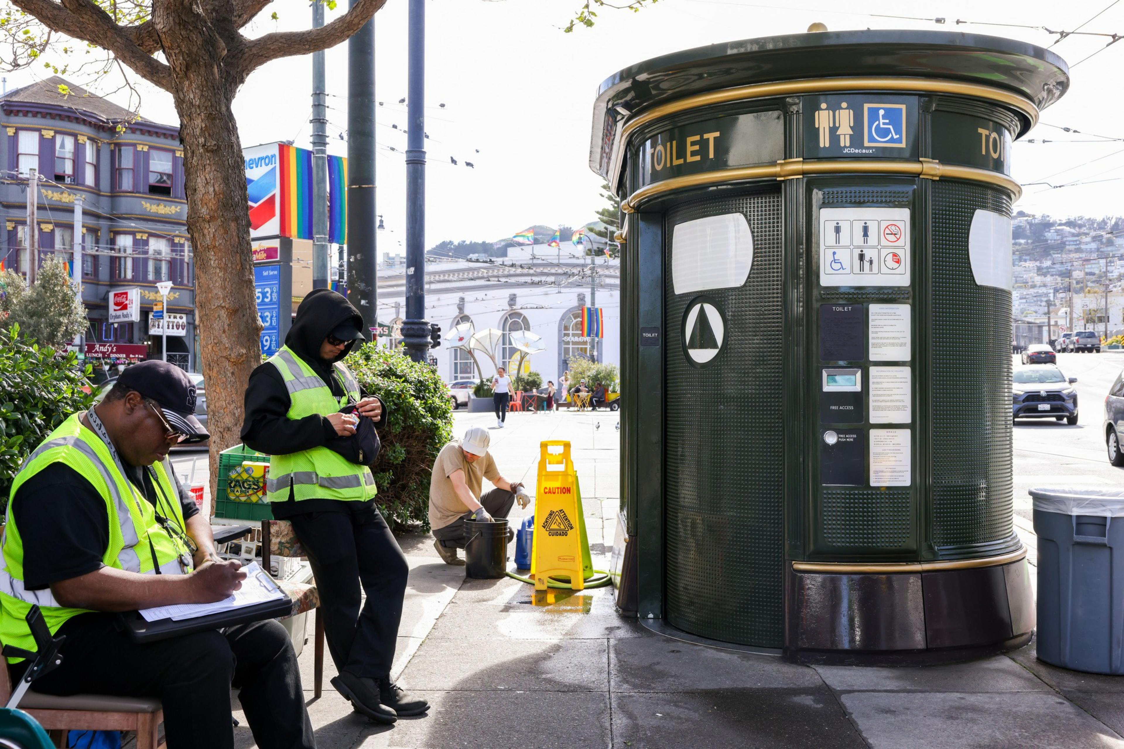 Two people in yellow vests stand beside a large public toilet. One is writing, and the other is eating. A cleaner works nearby with a caution sign.