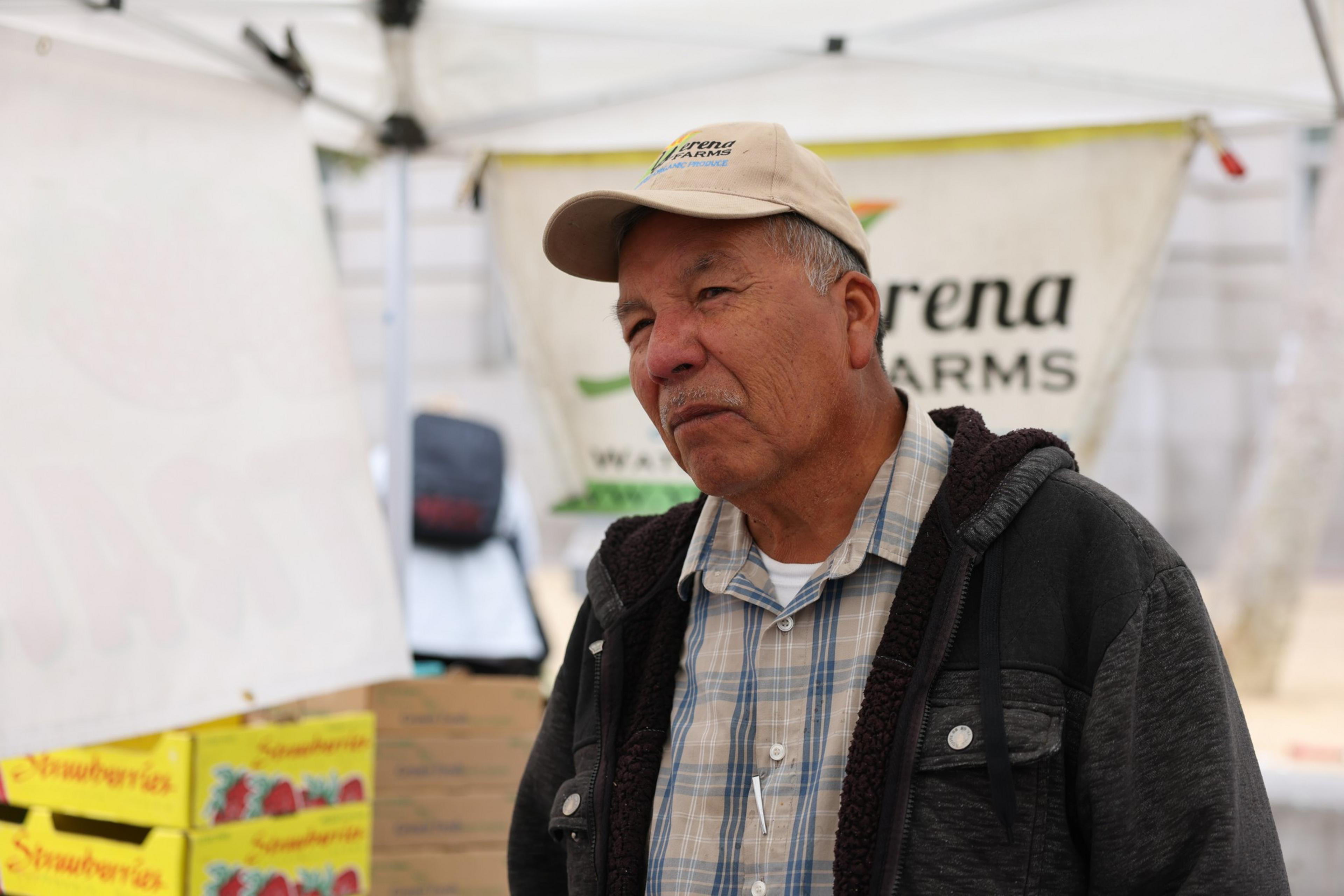 An older man wearing a cap and jacket stands in front of a tent with farm labels.