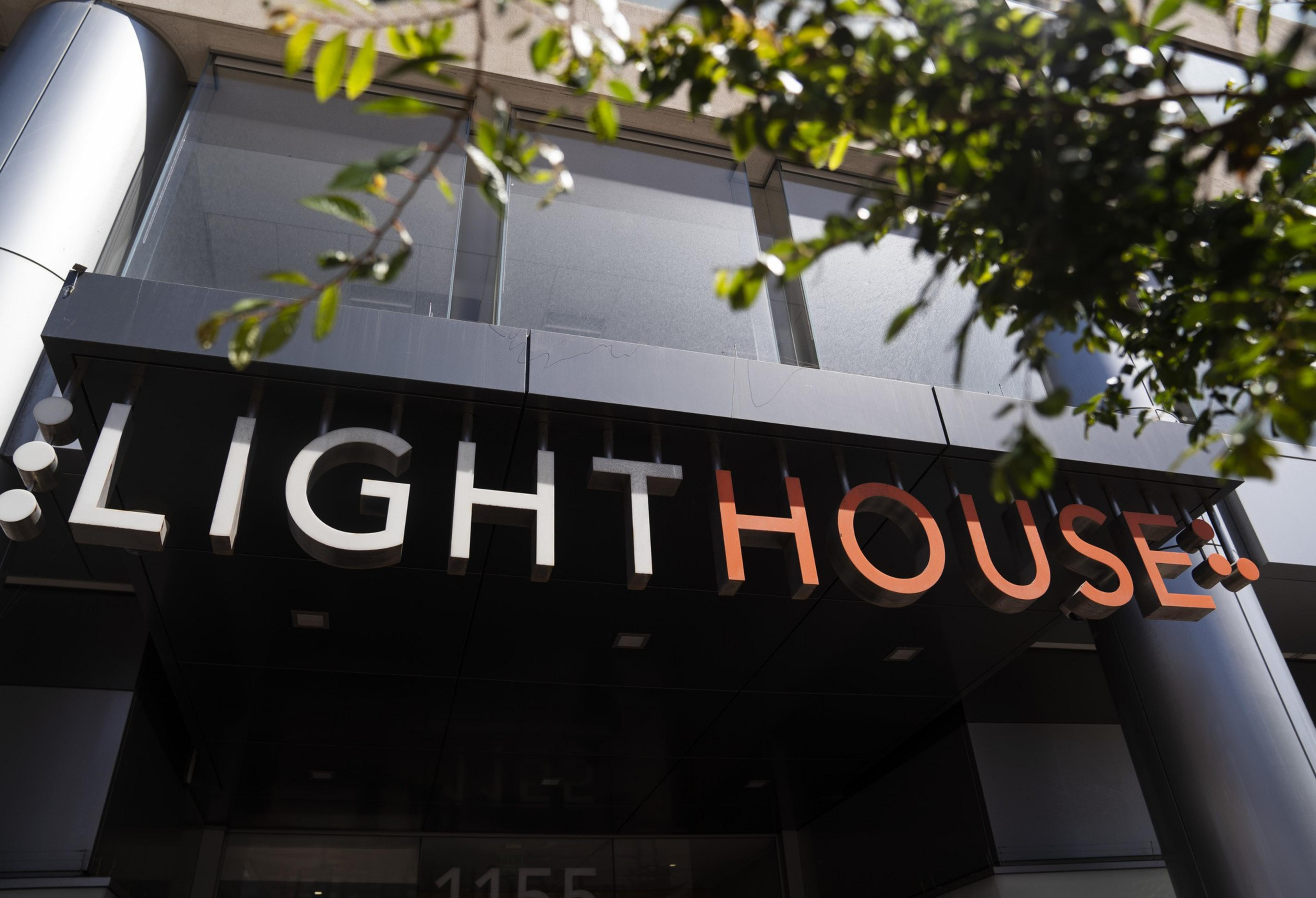 A dark building entrance is pictured with a sign reading &quot;LIGHTHOUSE&quot; in bold white and orange letters. Part of the sign is obscured by greenery.