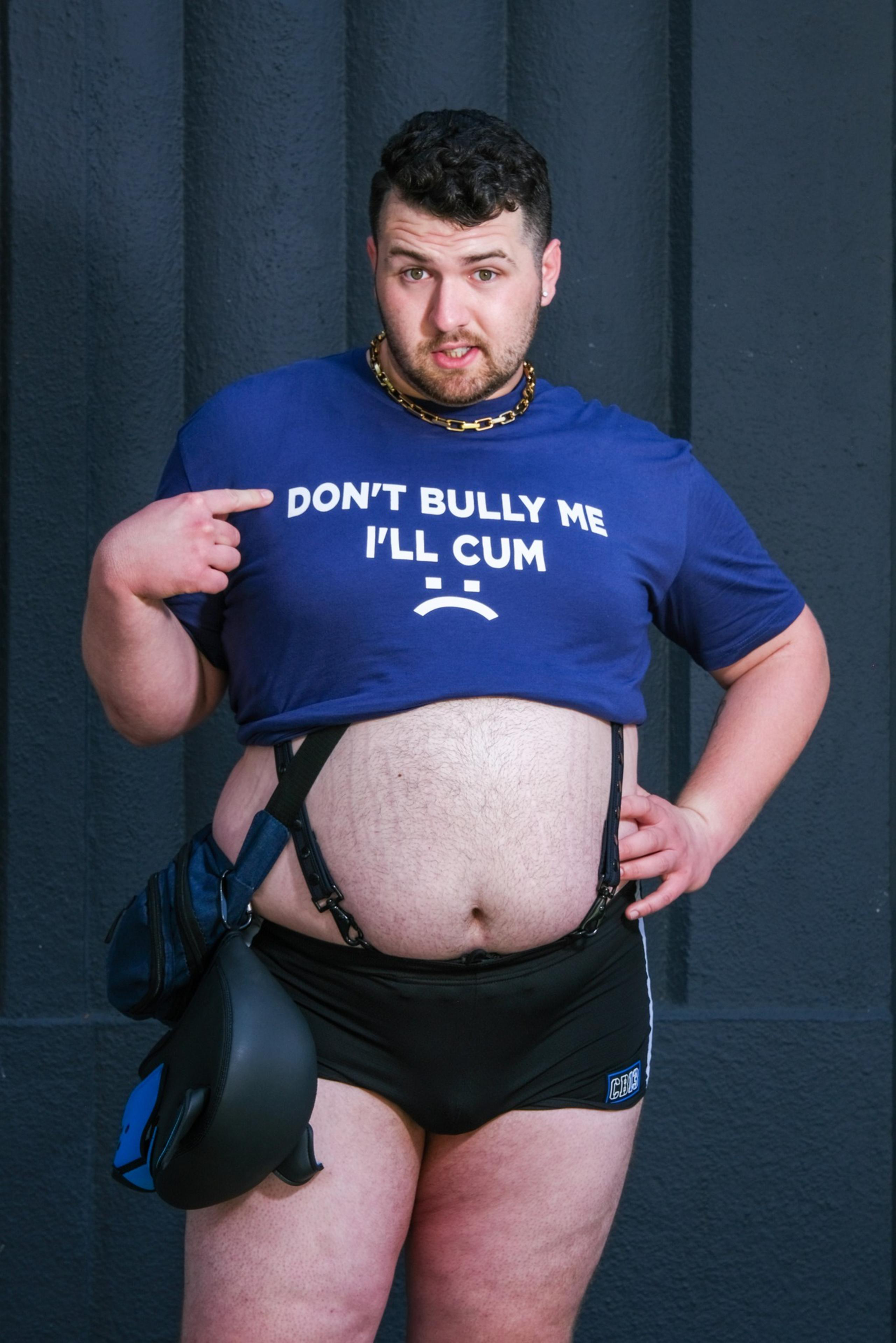A man stands against a dark background wearing a blue shirt that reads &quot;DON'T BULLY ME I'LL CUM,&quot; pointing at the text. He has a chain necklace and holds a helmet.