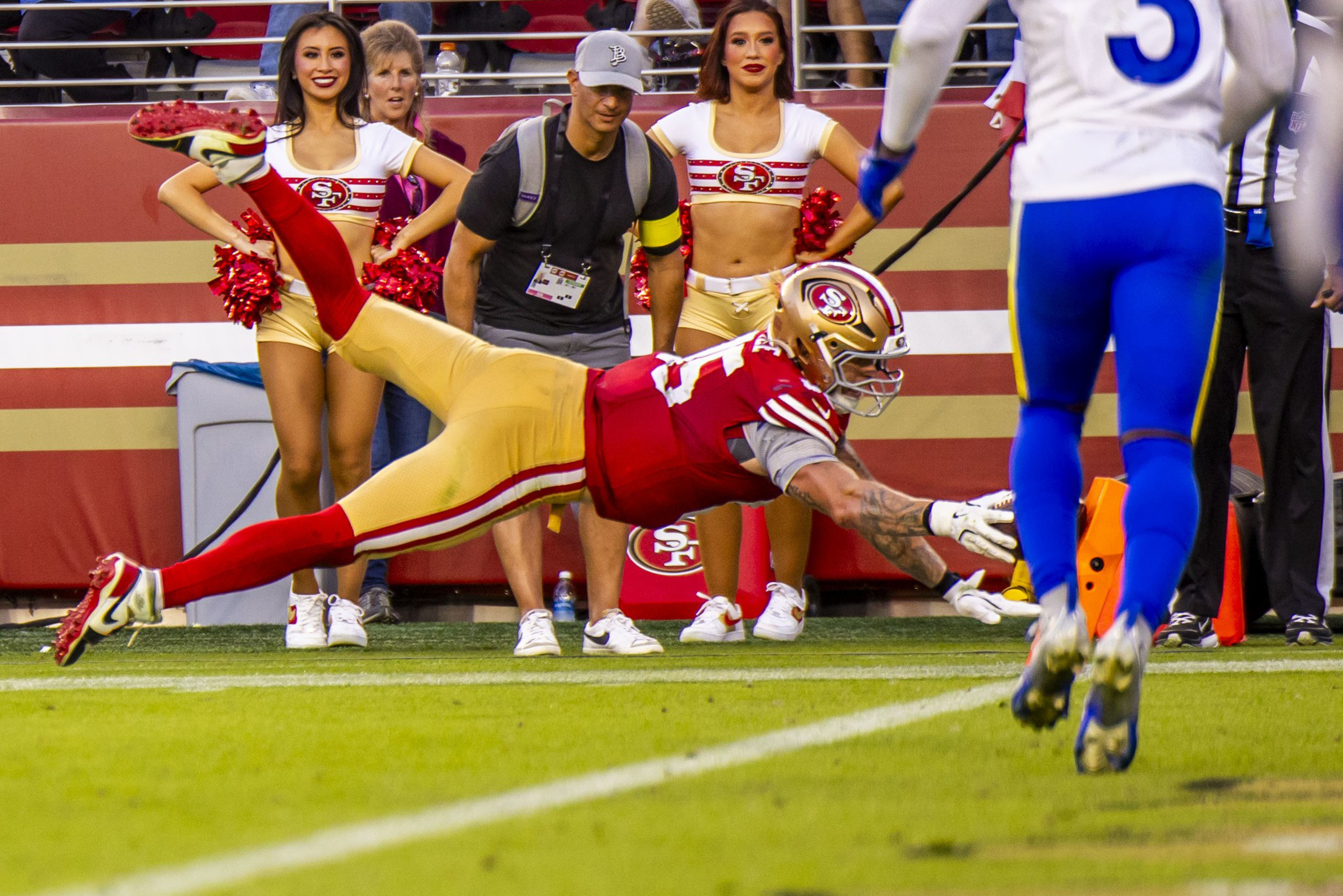 A football player in a red and gold uniform dives toward the sideline with outstretched arms as cheerleaders and staff watch nearby.